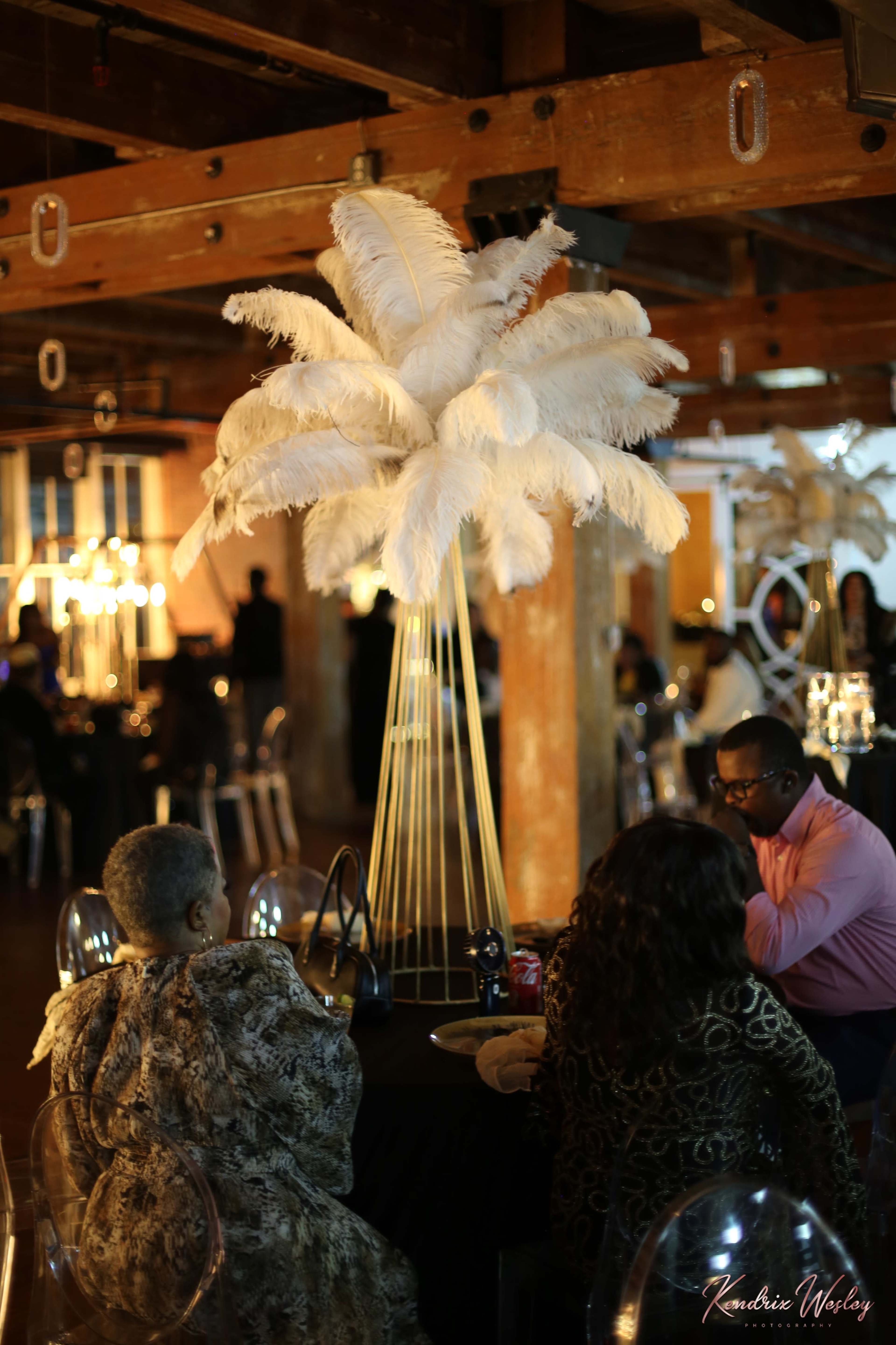 The image shows a decorated event space with elegant table settings and a large feather centerpiece.