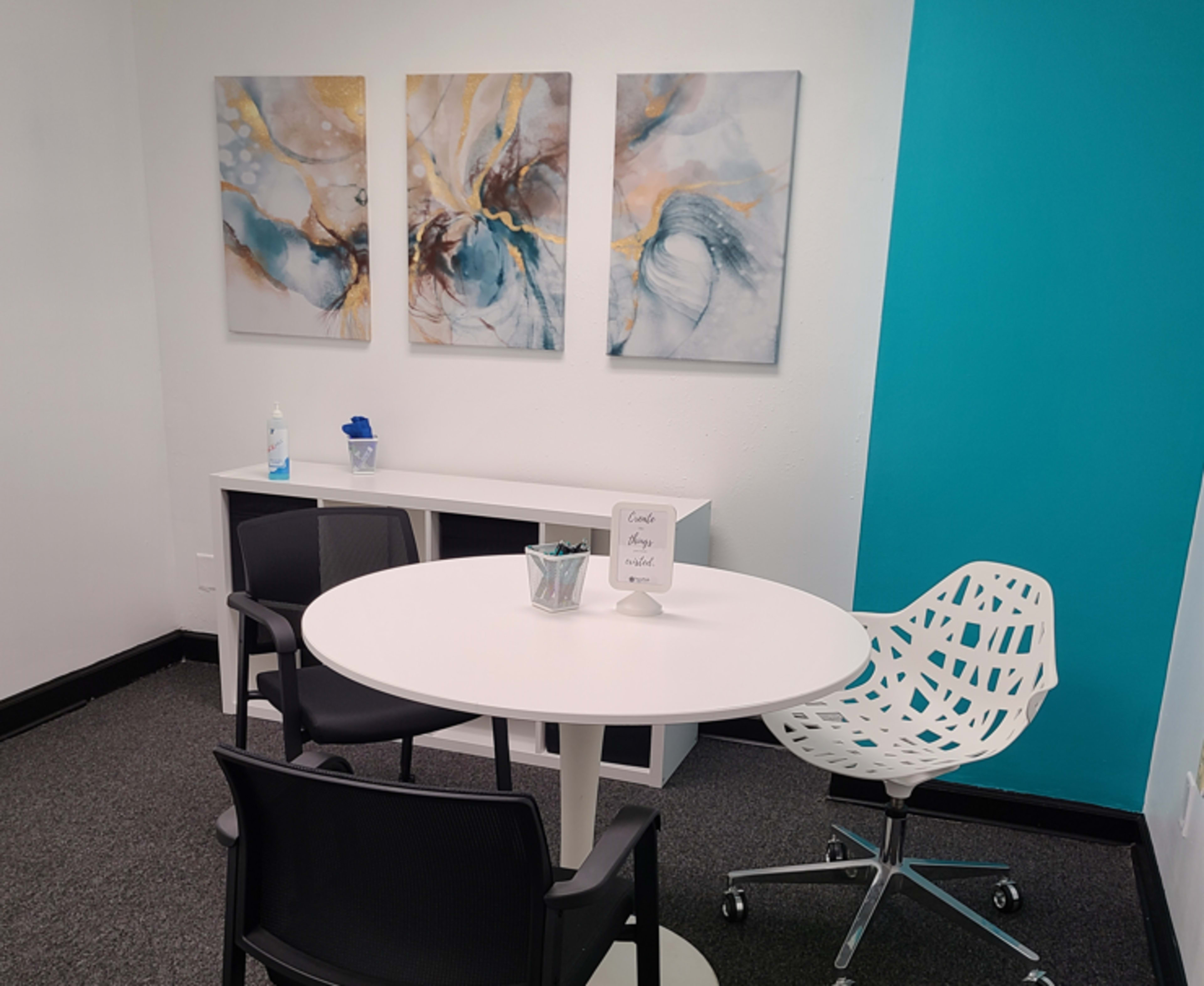A small office space with a round white table, two black chairs, a white decorative chair, a shelf with a blue container, and three abstract paintings on the wall.