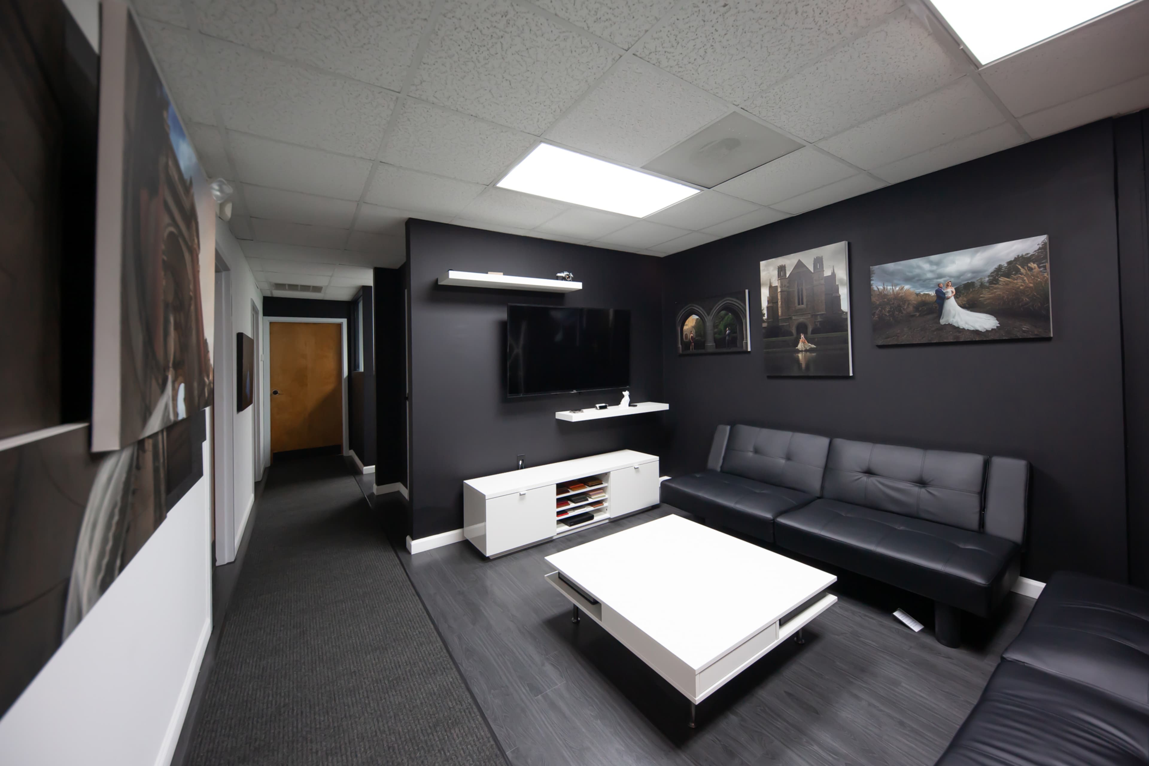 A modern office space with dark walls, a black sectional sofa, a white coffee table, and a television mounted on the wall, along with framed pictures displayed.