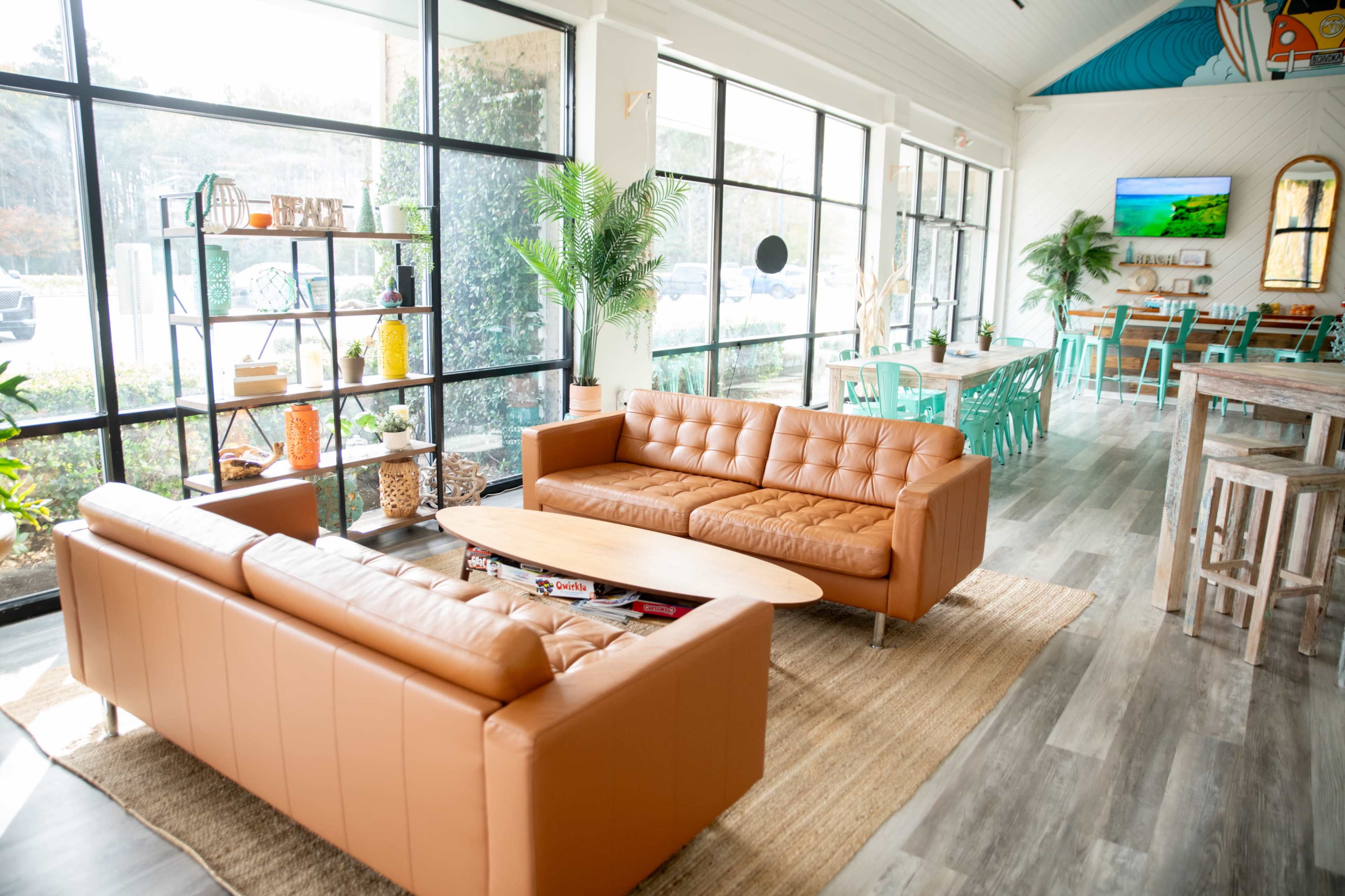 The image shows a bright and modern indoor space featuring two brown leather sofas, a long wooden table, and colorful chairs, with large windows allowing natural light to flood the room.