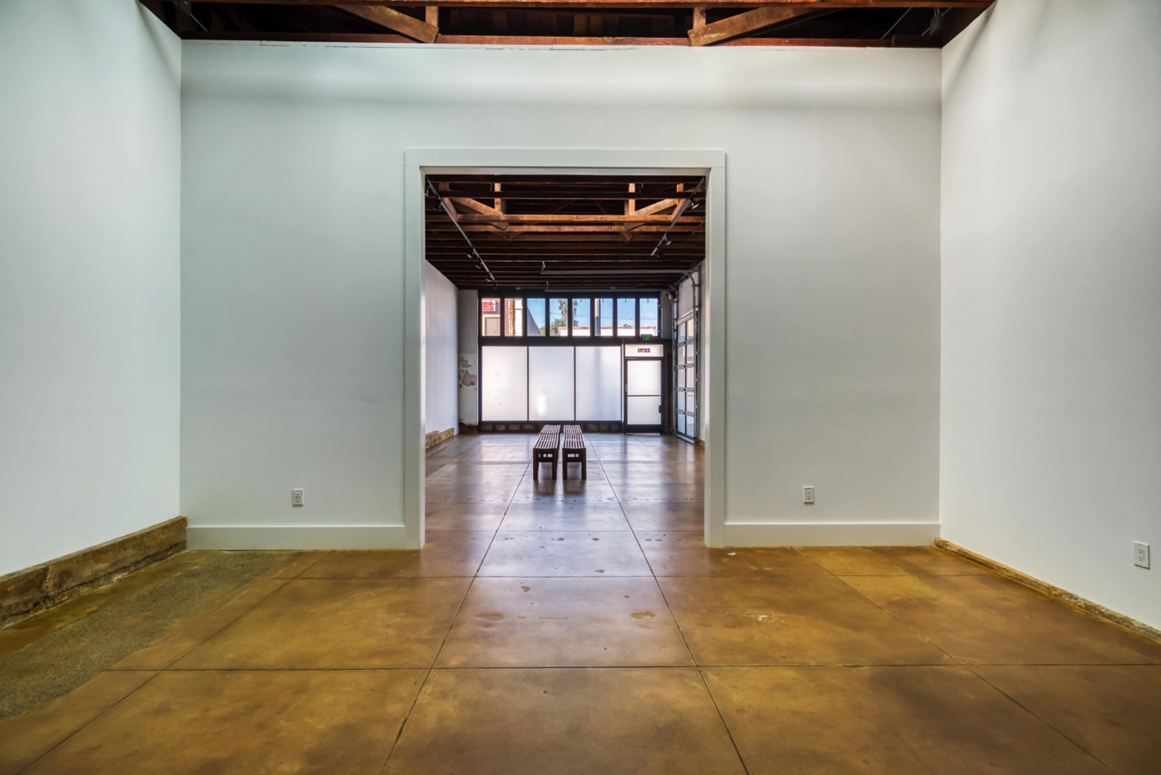 The image shows a minimalist interior space with a concrete floor, white walls, and exposed wooden beams, leading to another room with large windows.