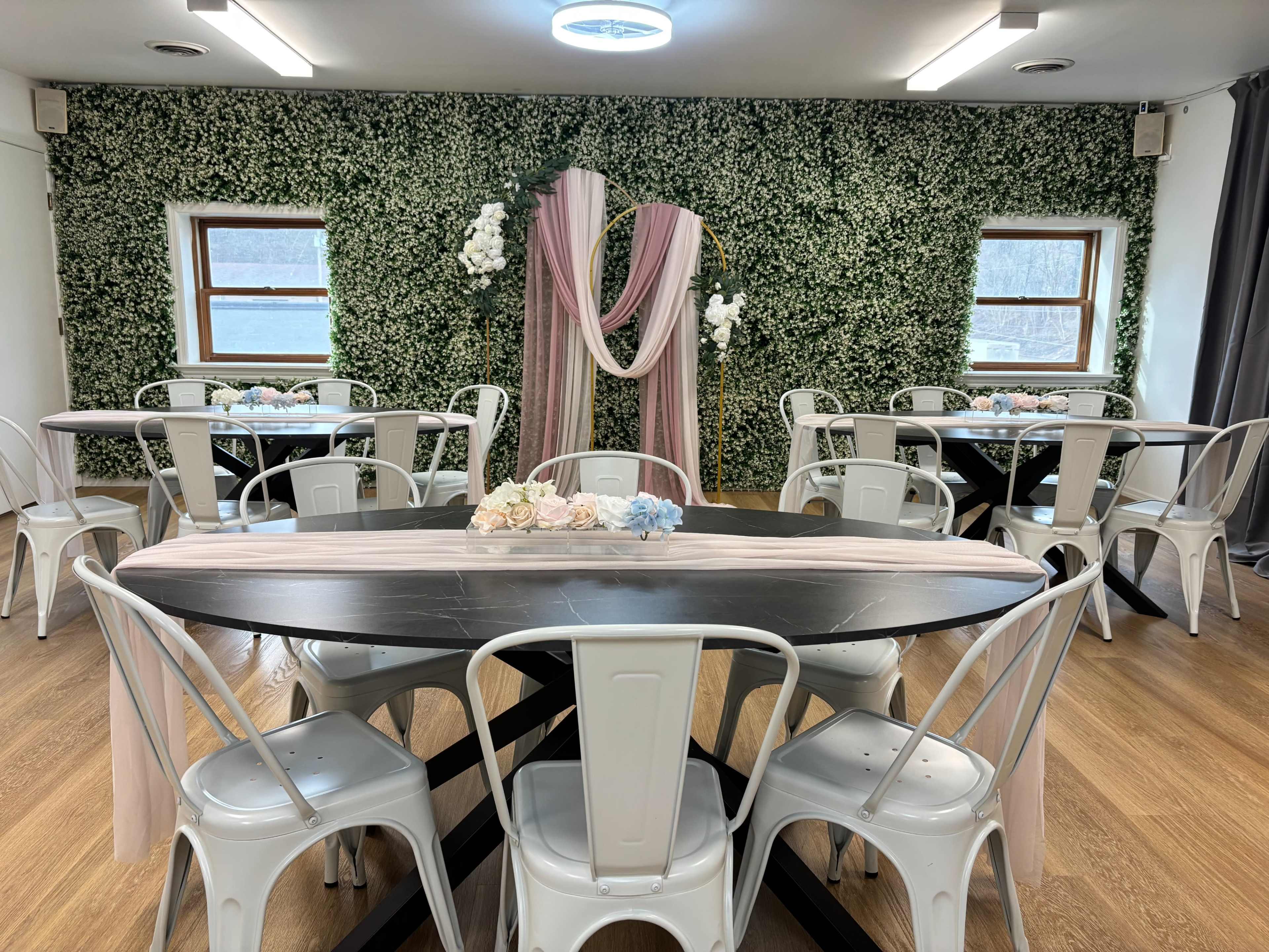 The image shows a spacious event room with several round tables surrounded by white metal chairs, decorated with a greenery wall and a floral arch at the front.