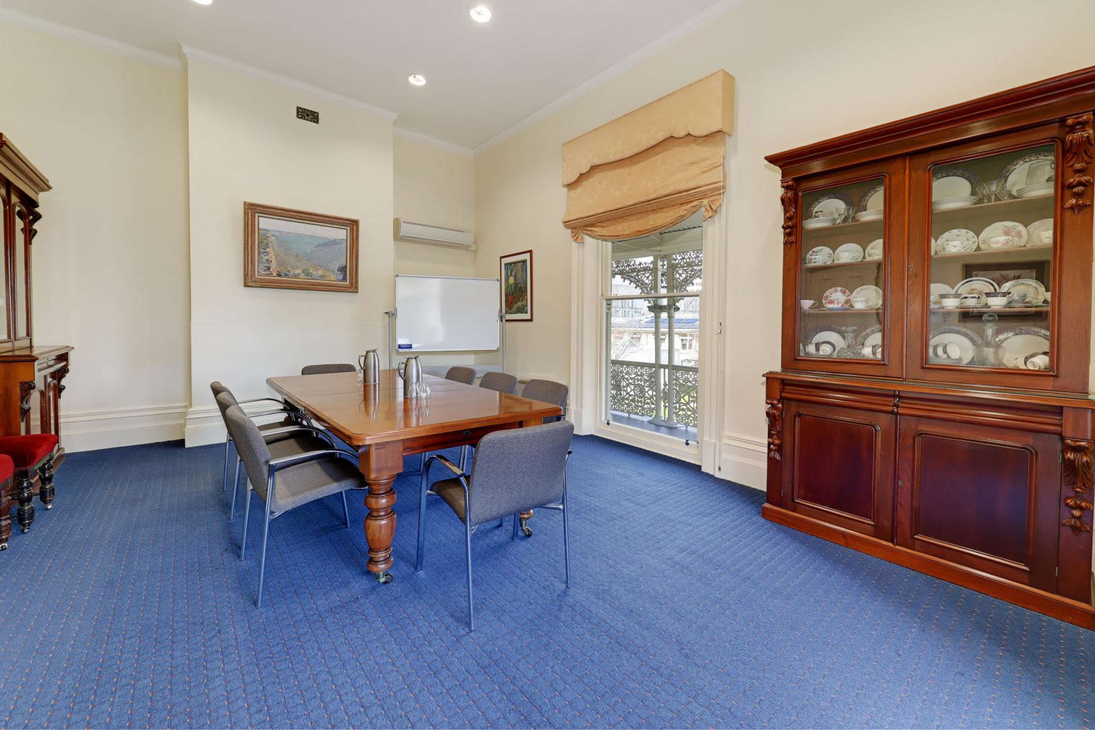 A conference room with a large wooden table, six gray chairs, and a cabinet displaying porcelain plates, illuminated by natural light from a window.