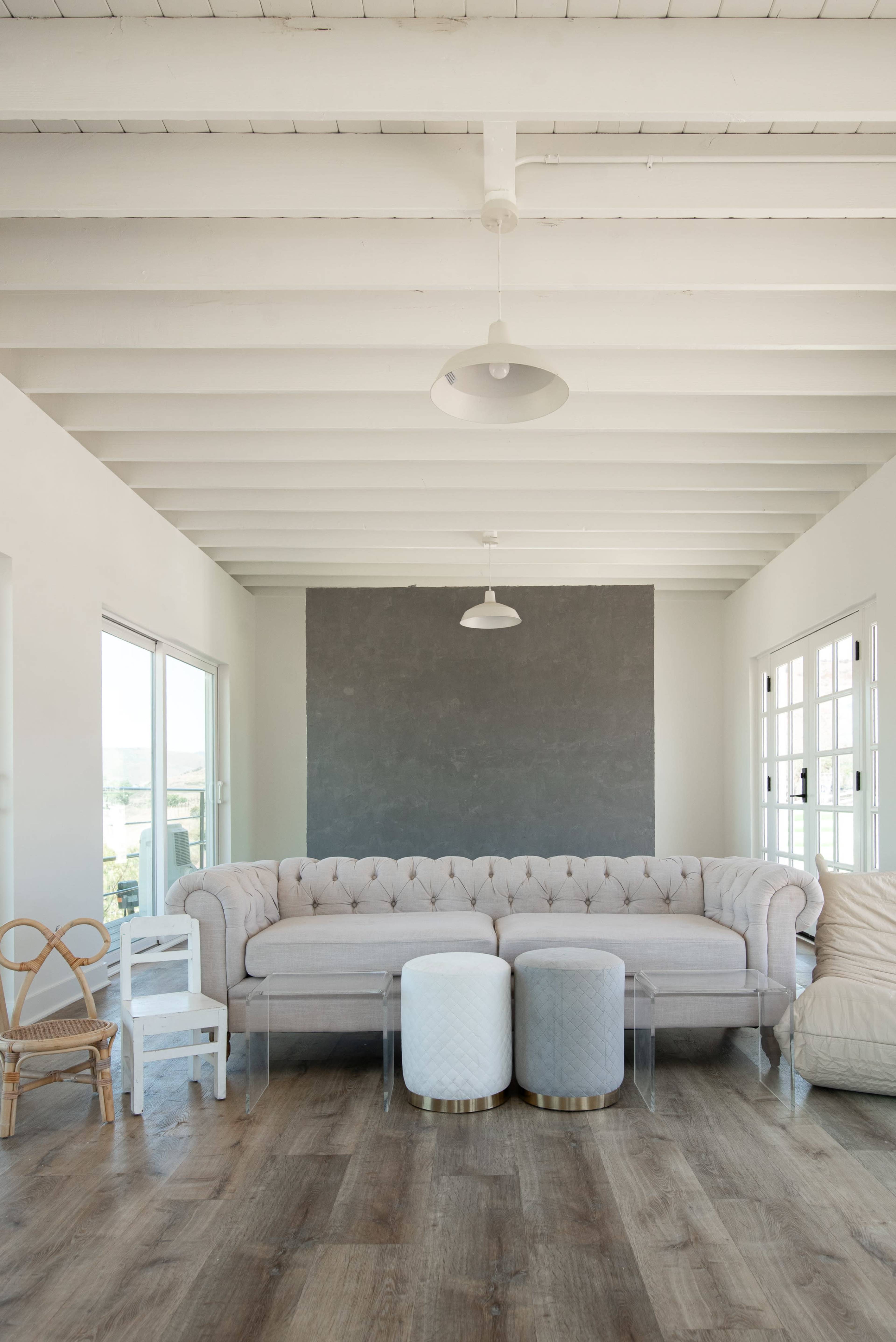 The image shows a modern living room featuring a light gray tufted sectional sofa, two round side tables, and a mix of seating options against a backdrop of white walls and exposed wooden beams.