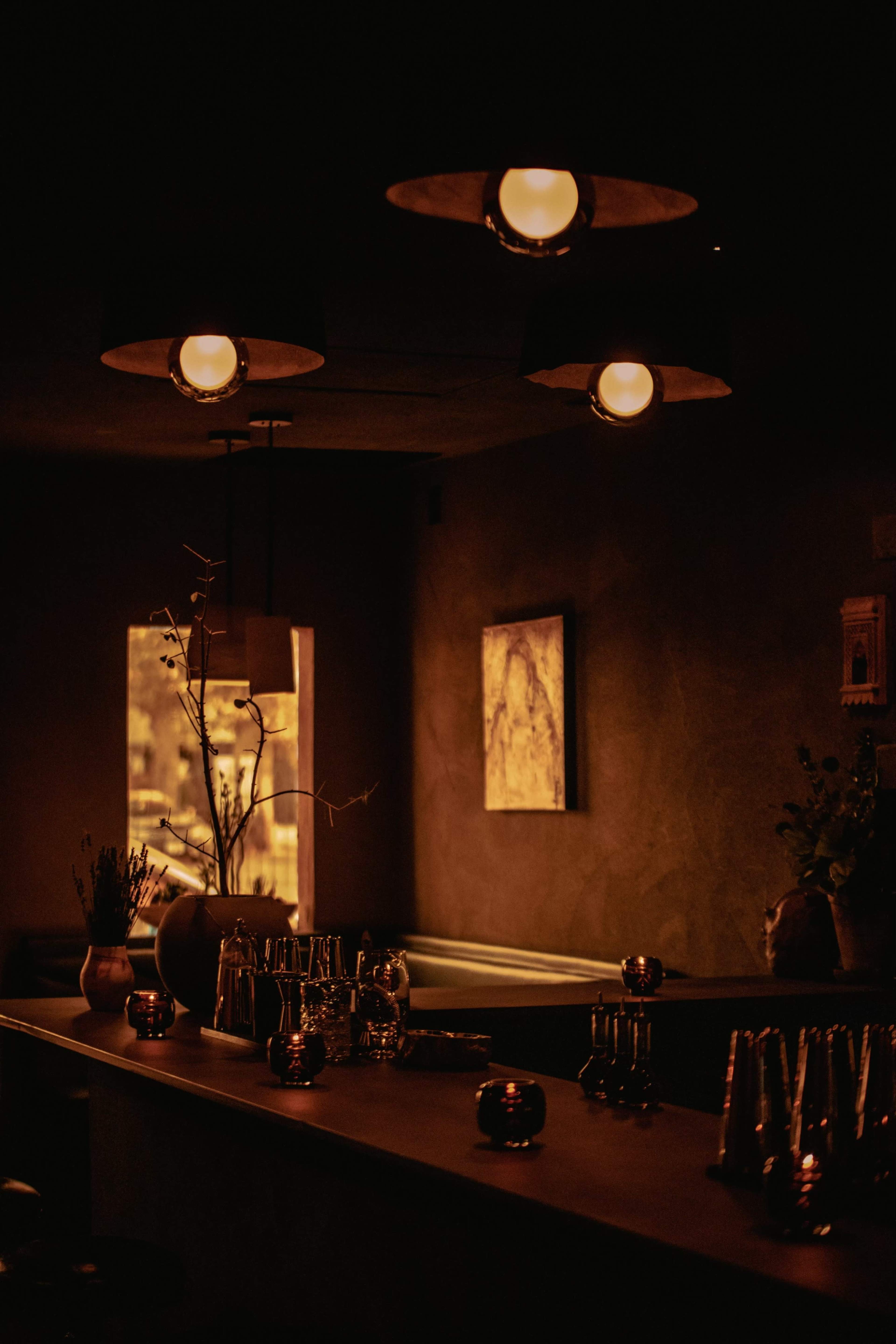 A dimly lit bar area with warm lighting, decorated with a few metallic accents and a large window illuminated from outside.