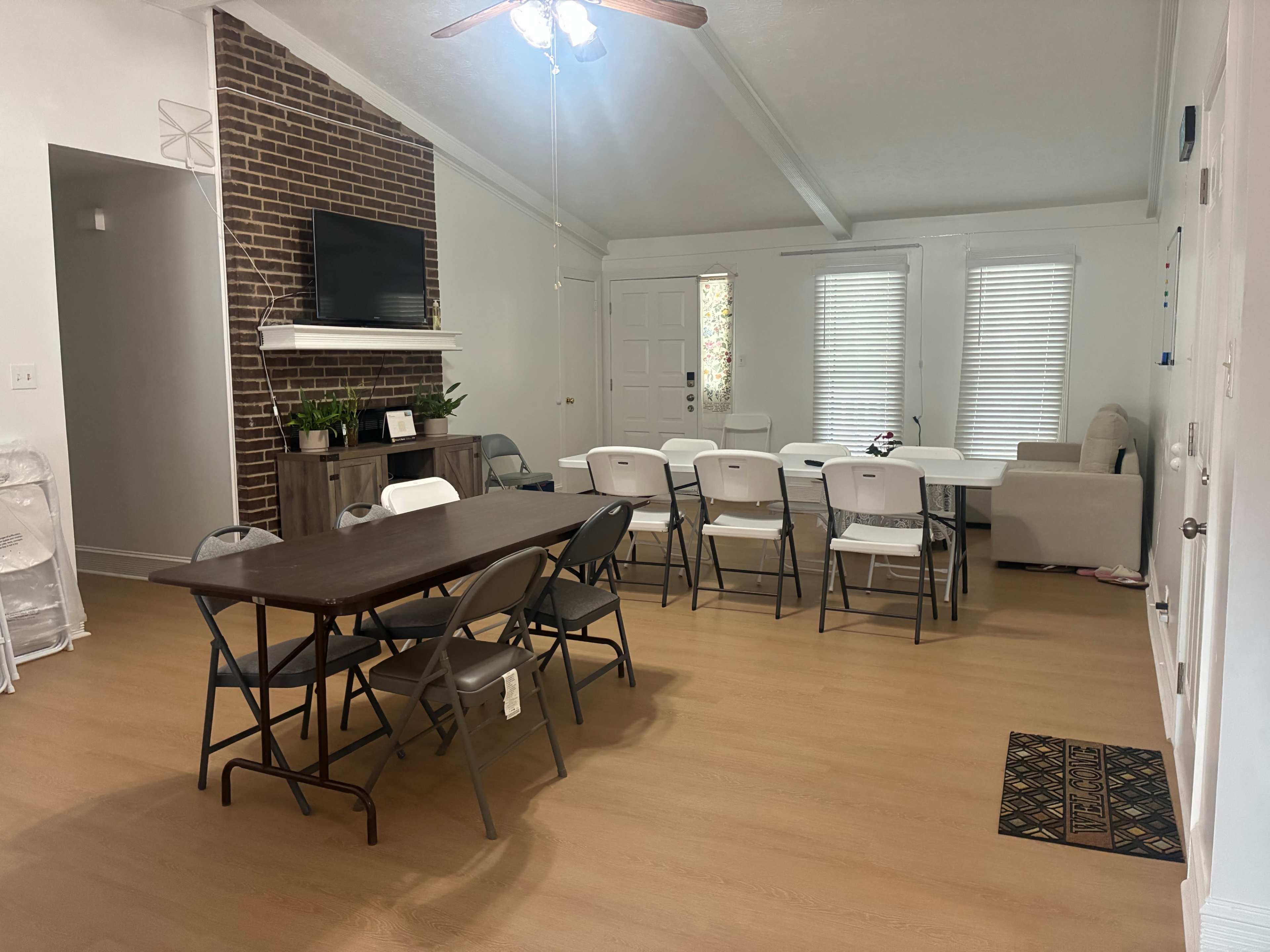 The image depicts a spacious room with a dining table and several folding chairs, a brick accent wall with a mounted television, and large windows allowing natural light.