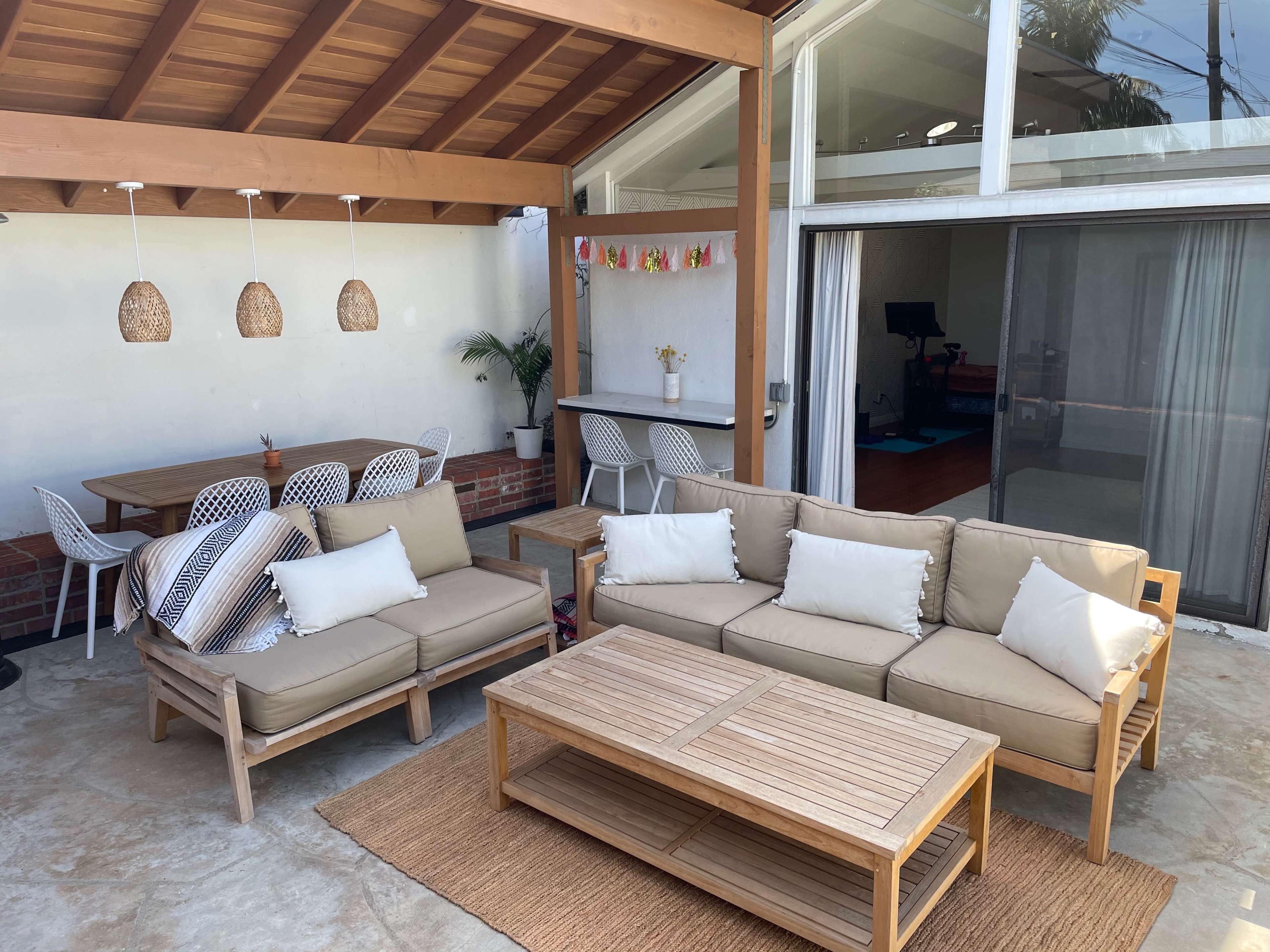 The image shows a well-furnished outdoor living space with a beige sectional sofa, a wooden coffee table, and a dining area with white chairs under a wooden ceiling.