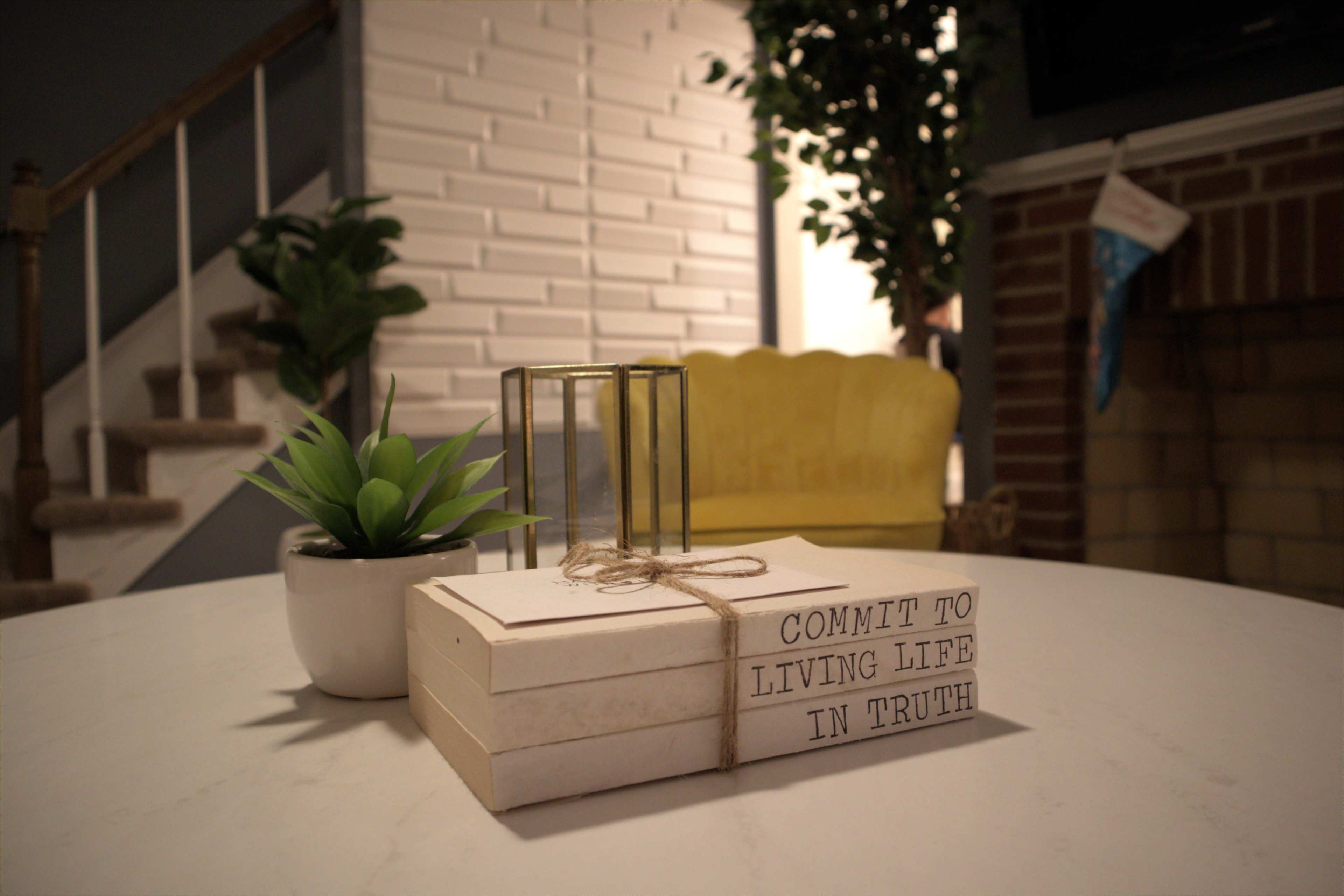 A decorative table displays a stack of books with the words "COMMIT TO LIVING LIFE IN TRUTH," a small potted plant, and a geometric terrarium, with a yellow chair and a brick wall in the background.