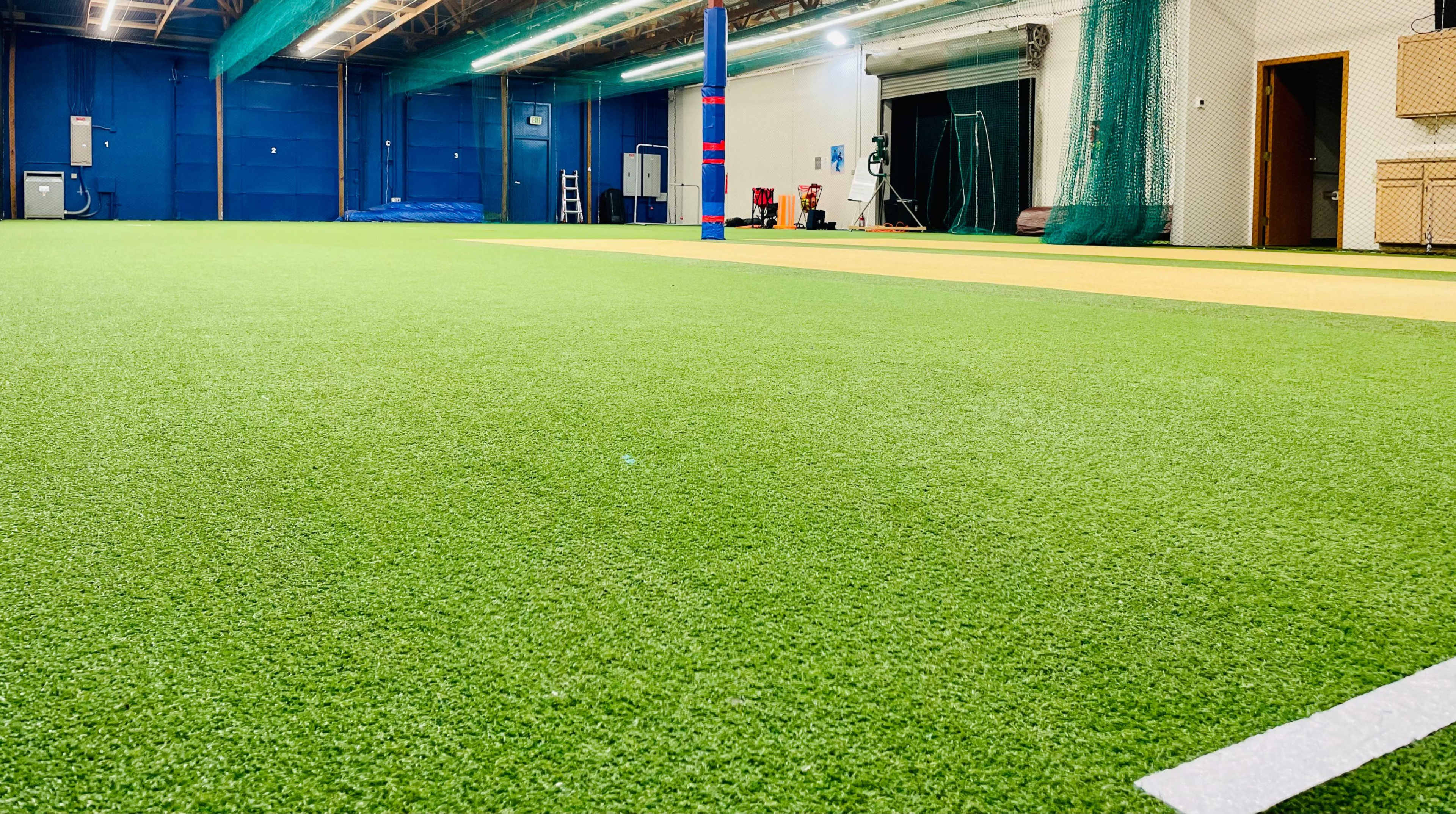 The image shows an indoor sports facility featuring artificial turf, with a blue wall in the background and various equipment along the sides.