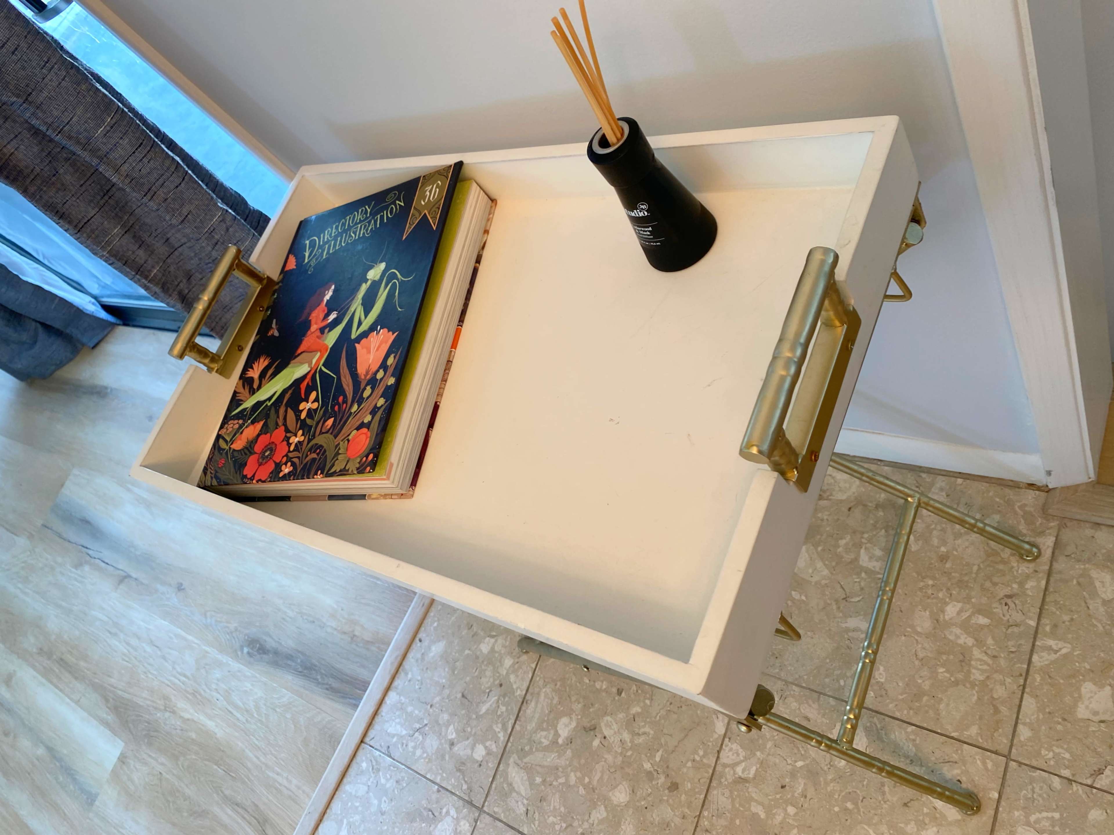 A white tray table with brass handles holds a book and a black vase with decorative sticks, positioned beside a window.