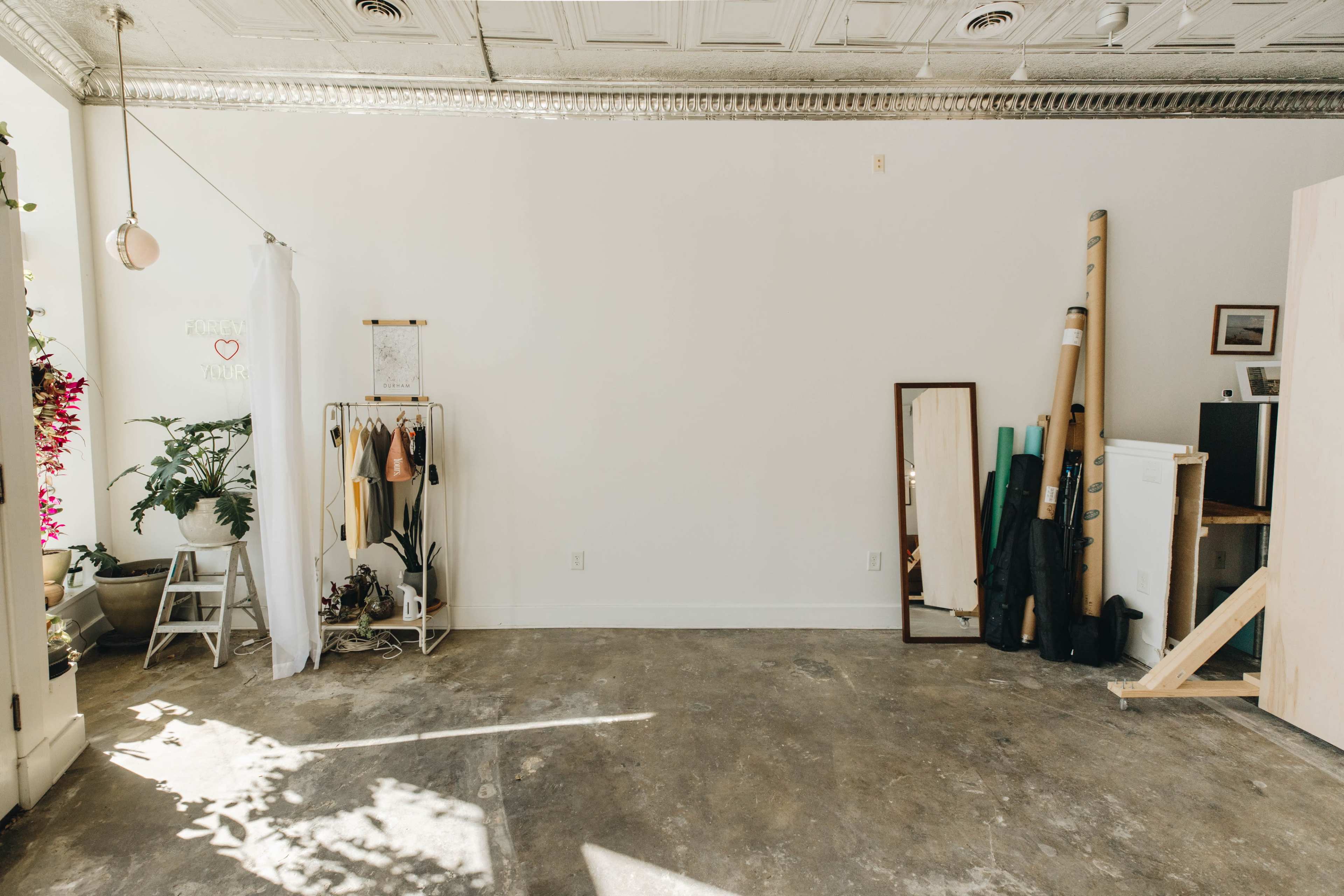 A spacious, minimally decorated room with concrete flooring, a mirror, a clothing rack, and various objects leaning against the wall.