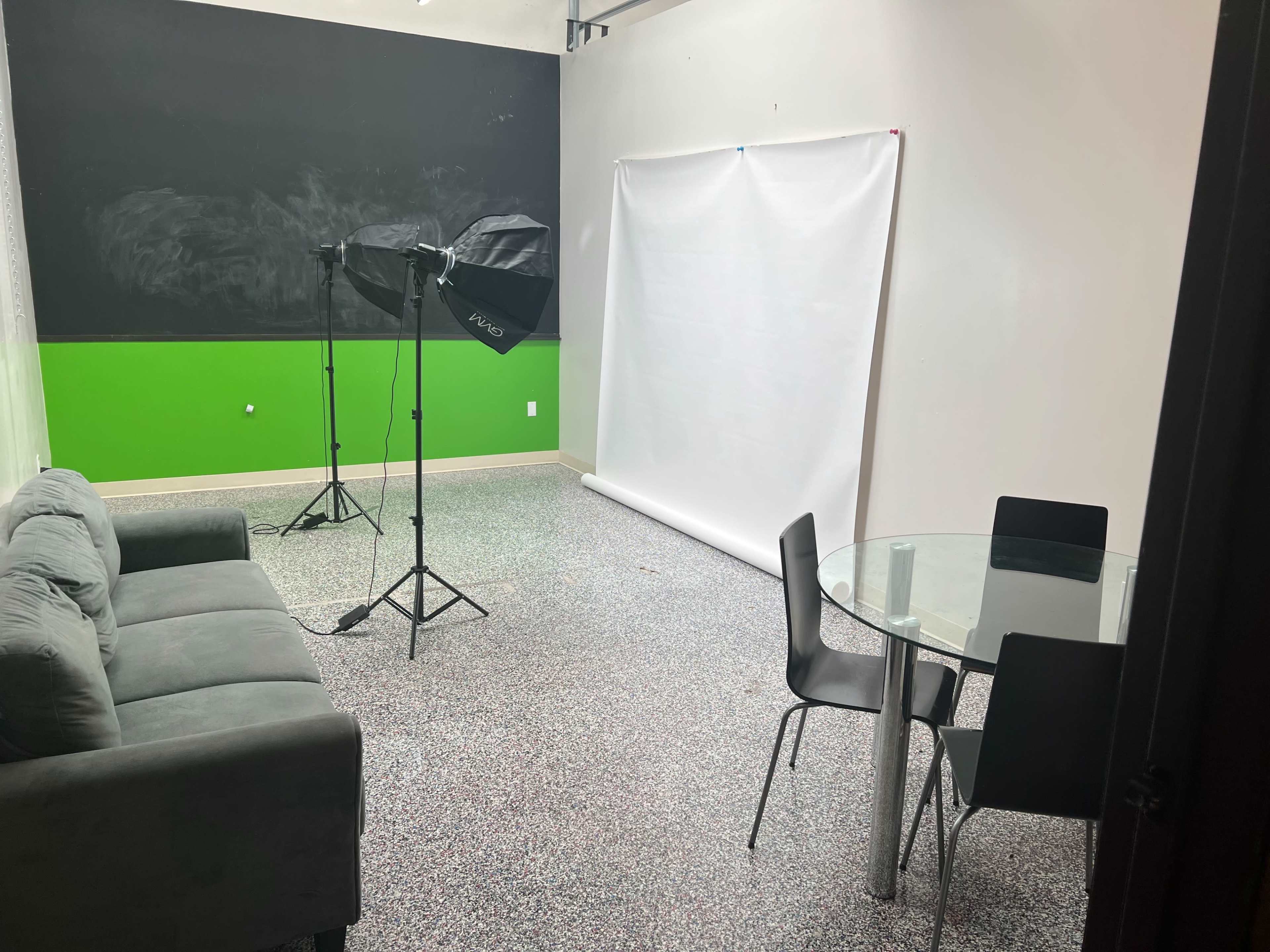 A small studio space with a gray couch, a round glass table with four black chairs, and lighting equipment set up near a white backdrop against a green wall.