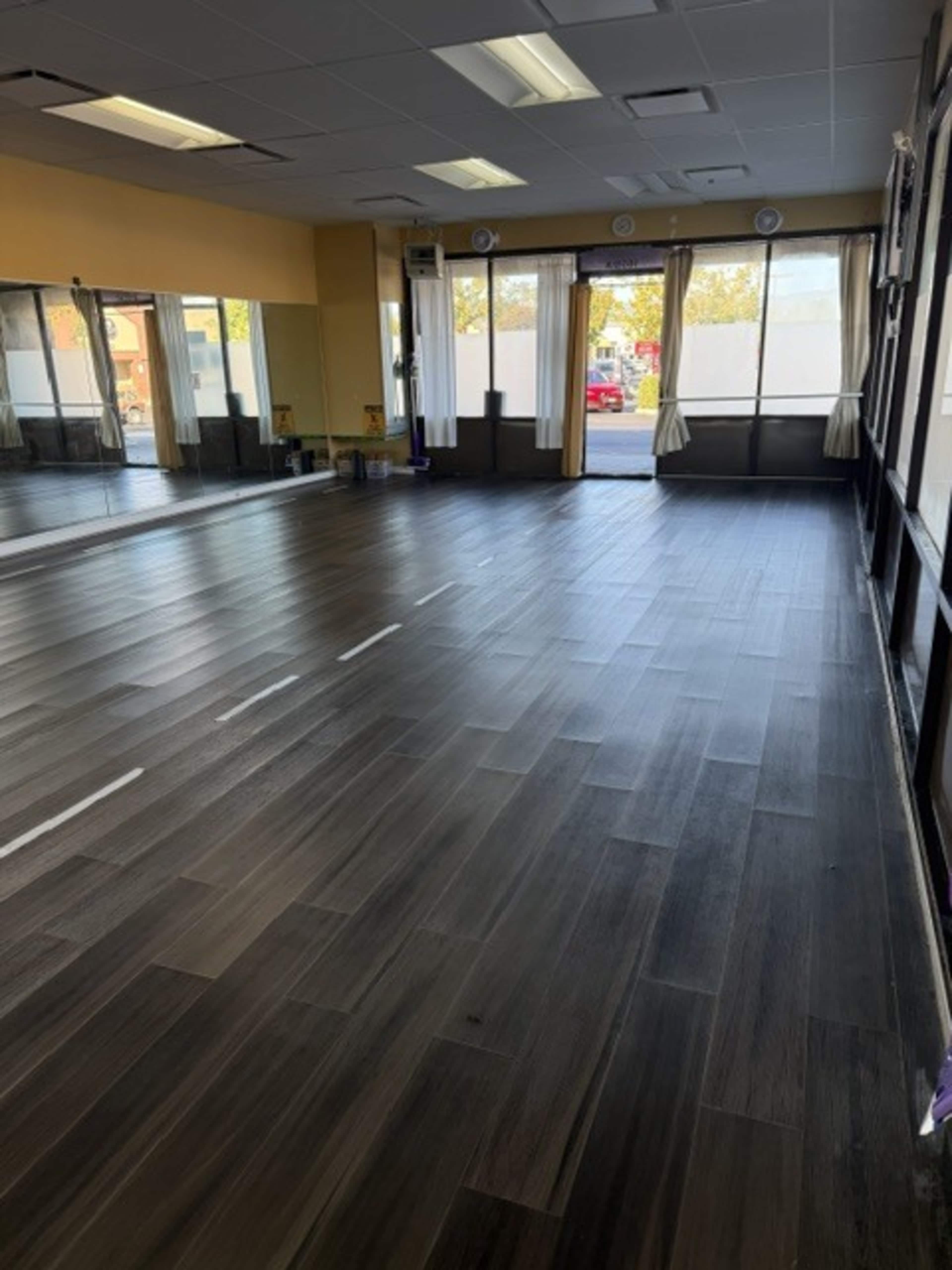 The image shows a spacious, brightly lit room with wooden flooring, large mirrors on one wall, and windows allowing natural light to enter.