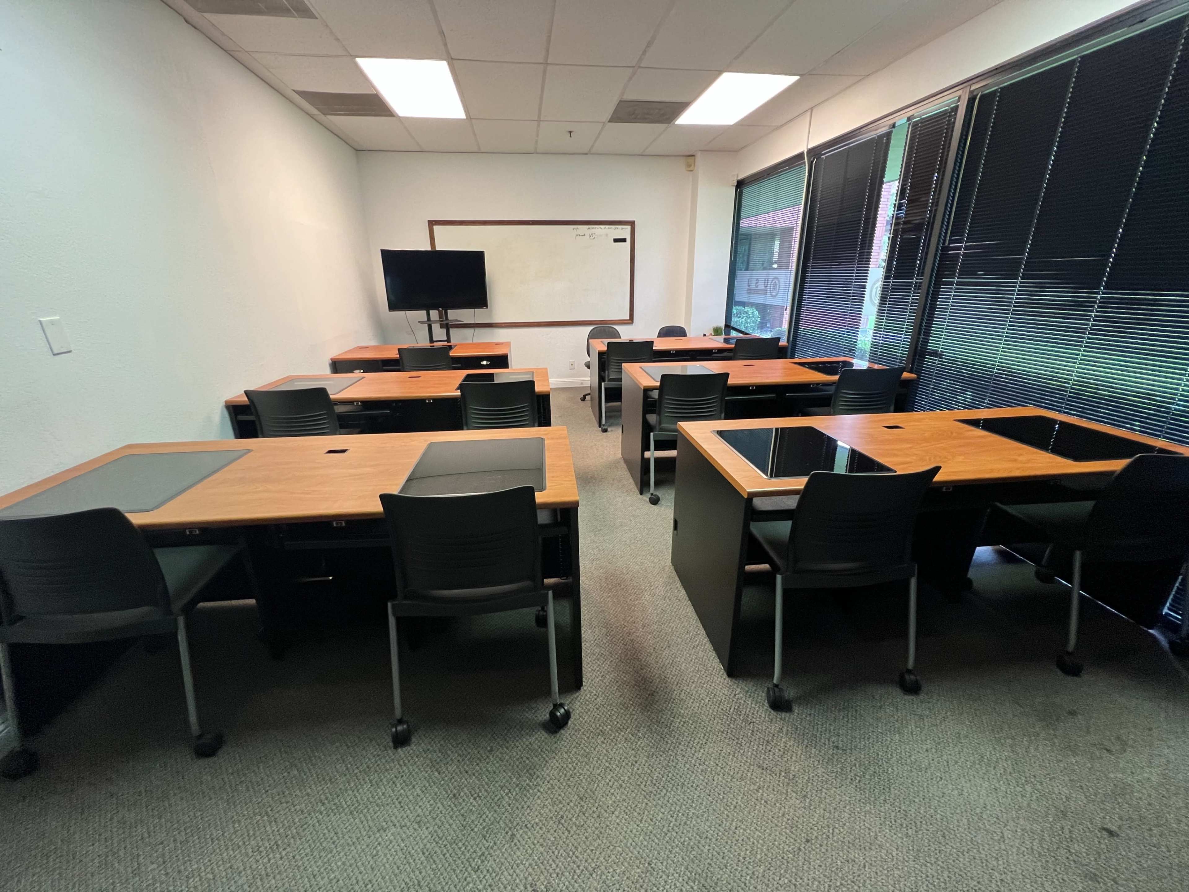 A classroom with multiple wooden desks arranged in rows, equipped with black chairs, and featuring a television and whiteboard at the front.
