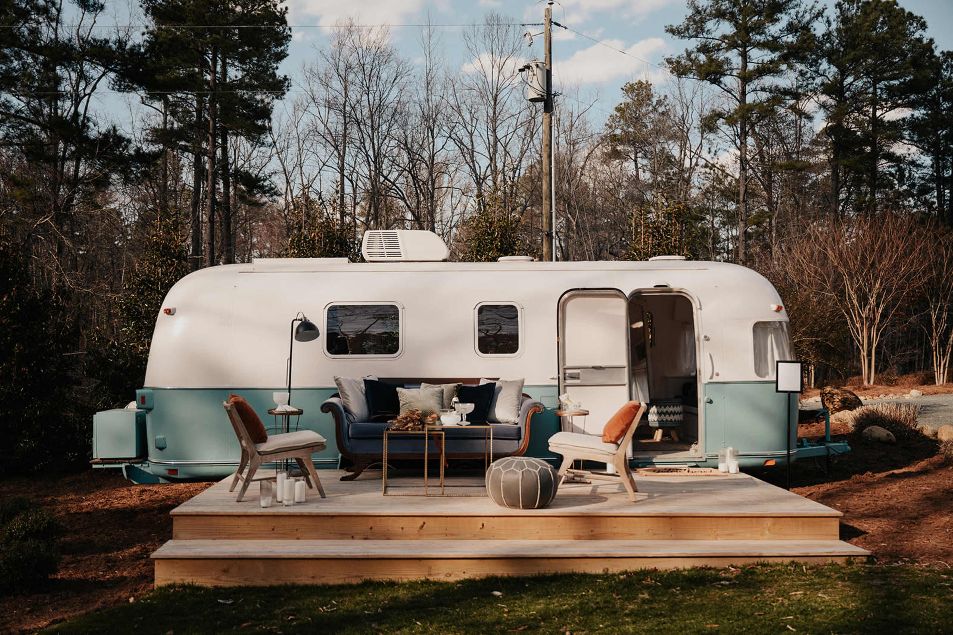 A vintage trailer is parked on a wooden deck in a wooded area, surrounded by trees.