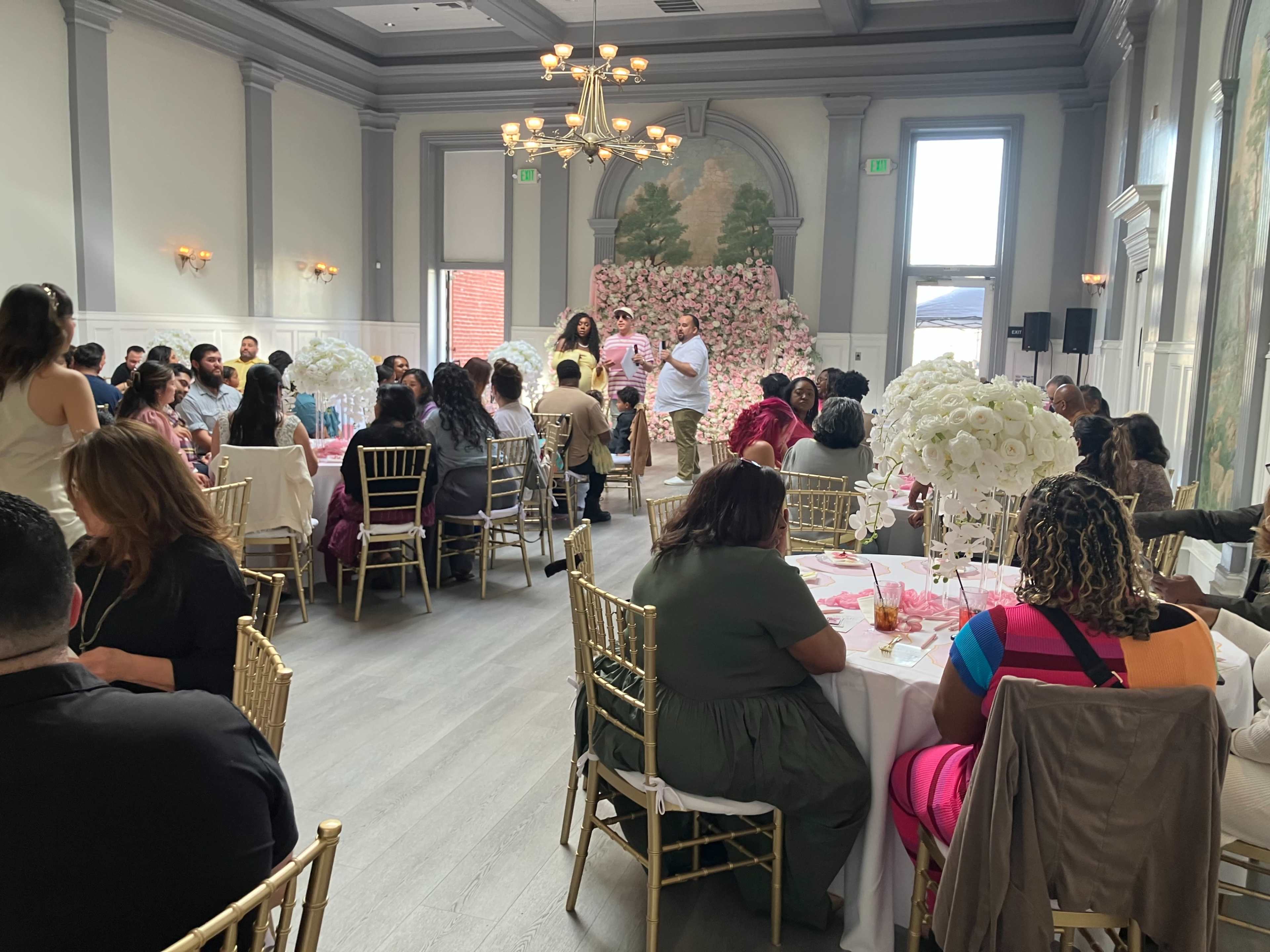 A large gathering takes place in a decorated event space, featuring guests seated at tables and a floral backdrop.