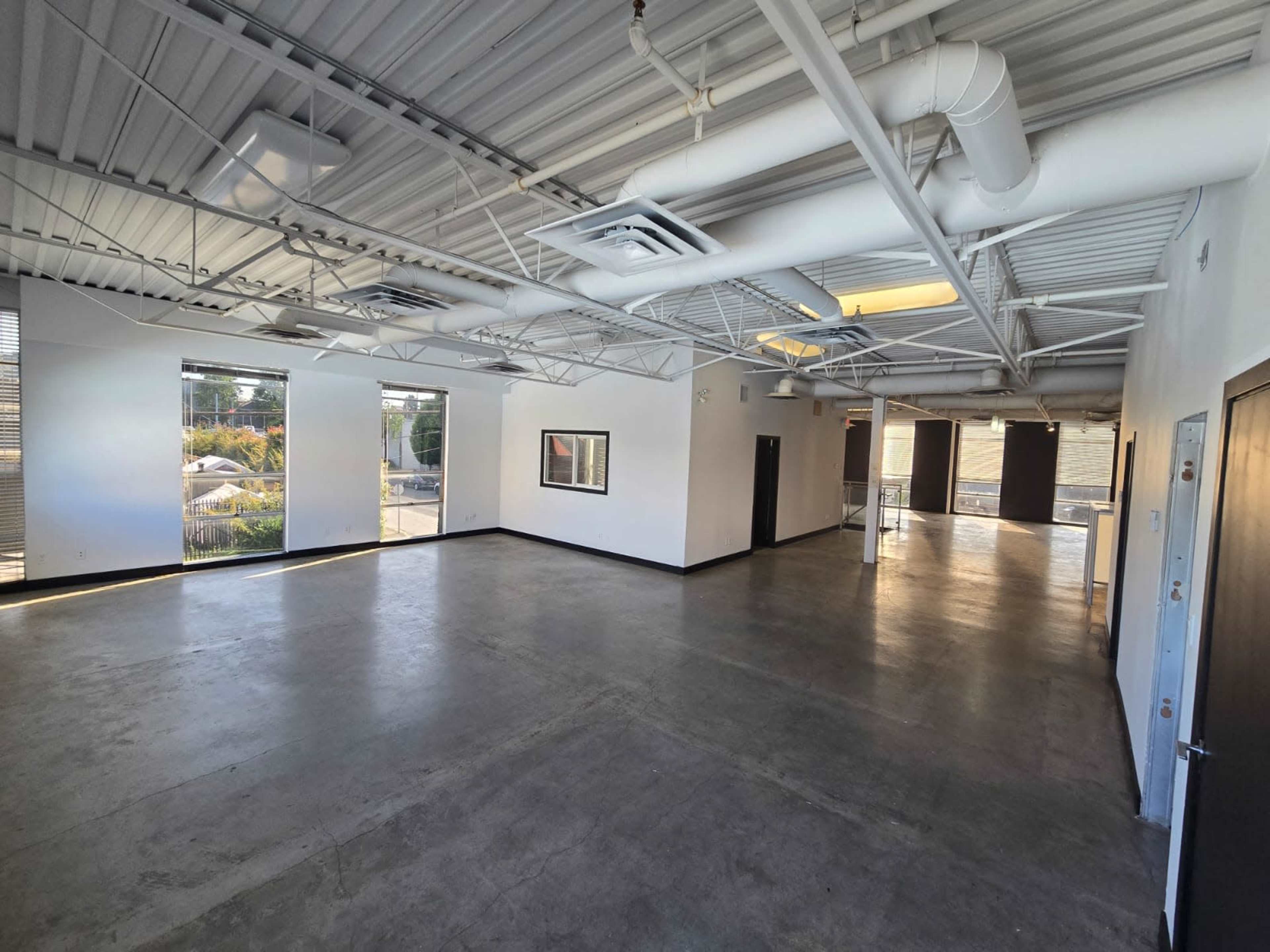 The image shows a large, empty room with concrete floors, white walls, and multiple windows allowing natural light to enter.