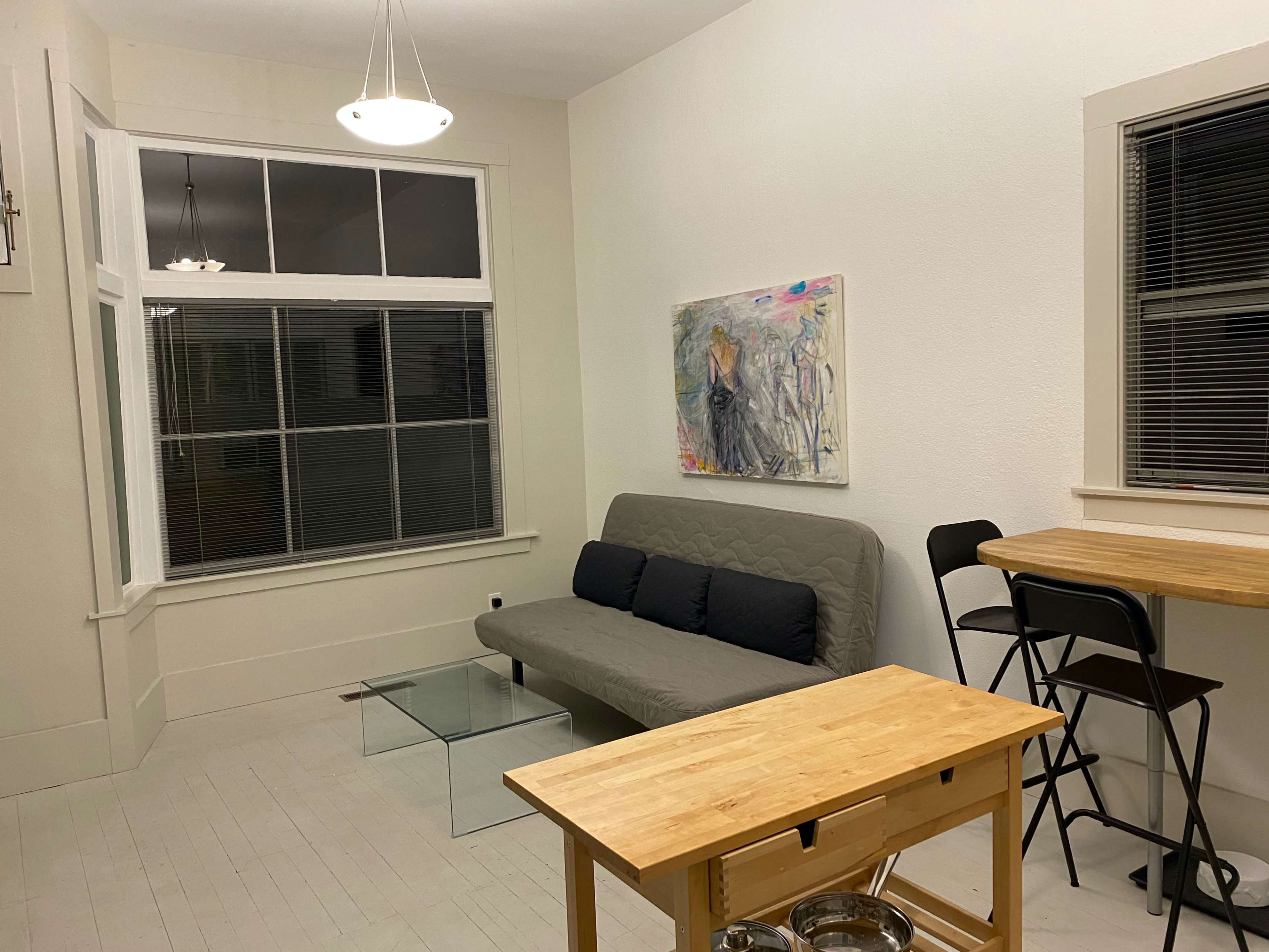 A well-lit room with a sofa bed, a glass coffee table, and a wooden dining table with high chairs, along with a large window and a piece of artwork on the wall.