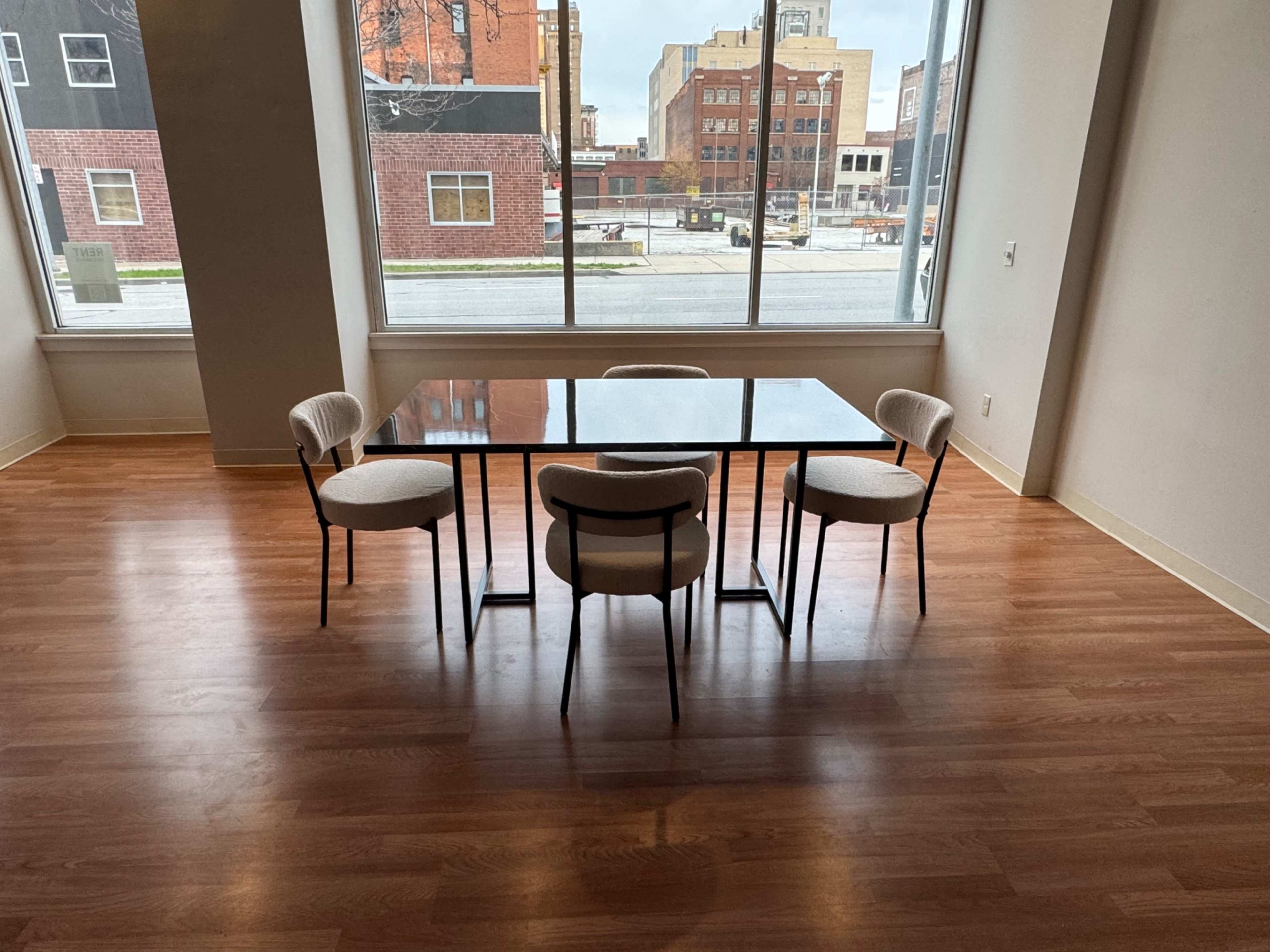 The image shows a simple dining table with four chairs, positioned in a well-lit room with large windows overlooking a cityscape.