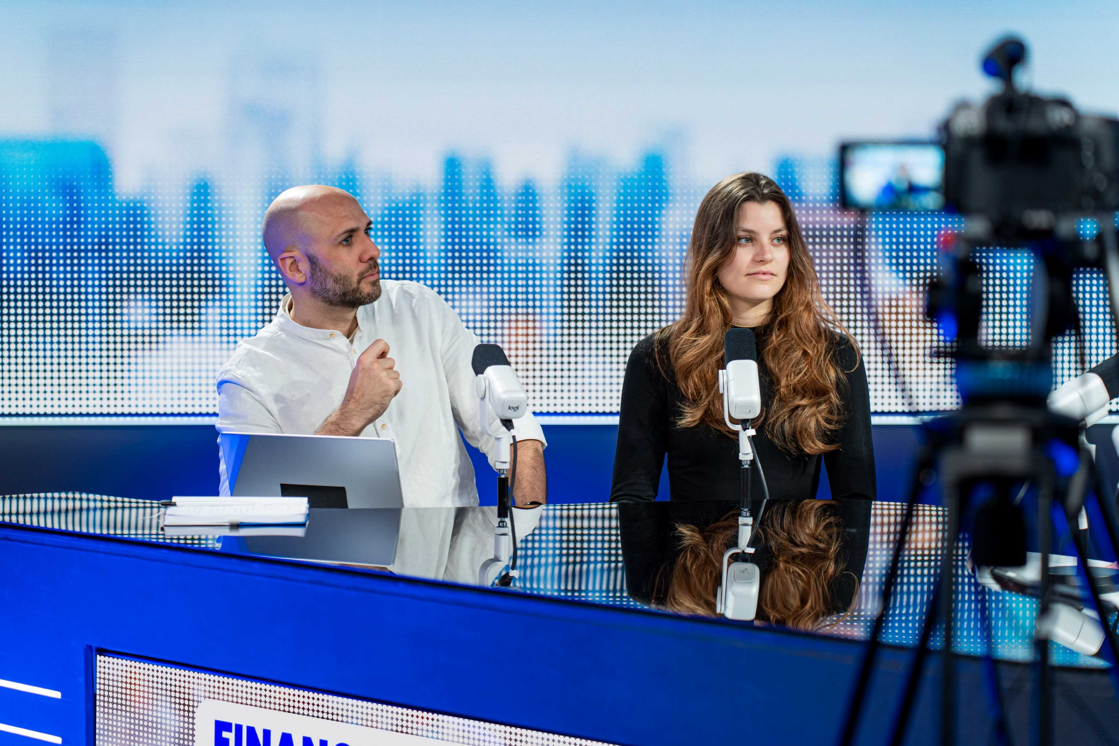 A man and a woman sit at a modern news broadcast studio desk, each facing a microphone while a camera captures the scene.