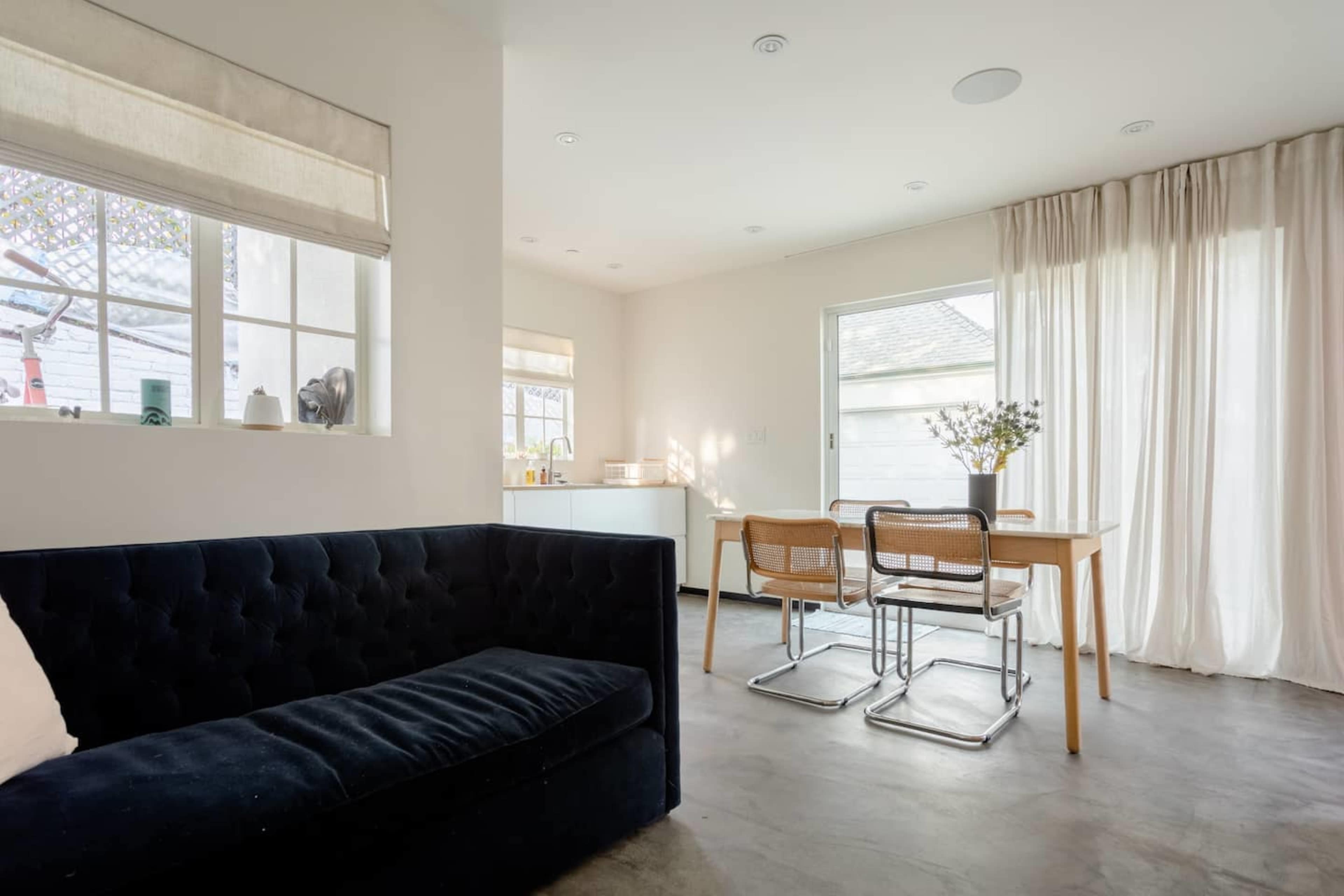 A modern living space features a dark sofa, a dining table with chairs, and large windows allowing natural light.