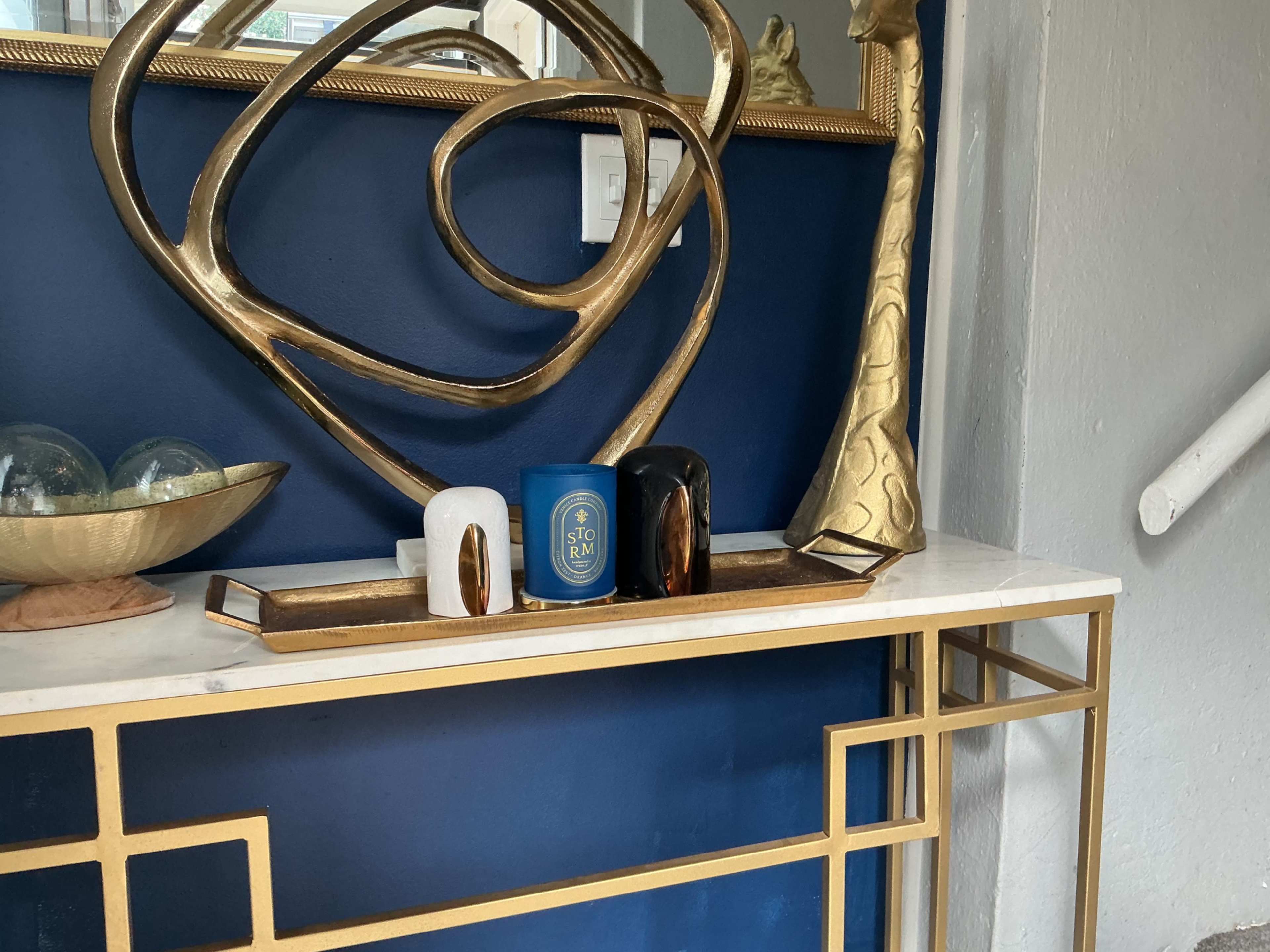 A console table with a marble top displays decorative items, including candles and an abstract gold sculpture, against a deep blue wall.