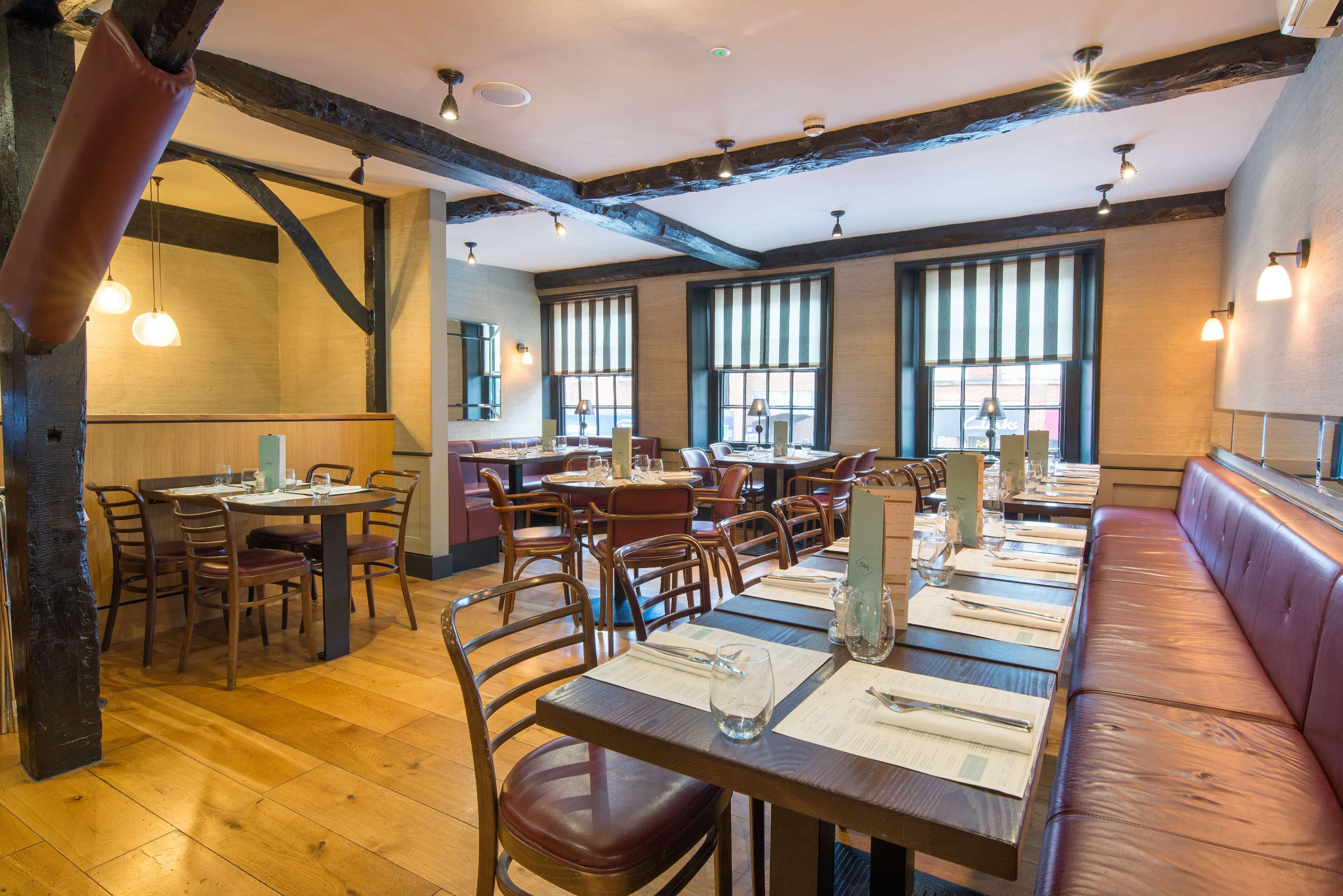 The image shows a warm, inviting restaurant interior featuring wooden beams, tables with metal chairs, and large windows with striped curtains.