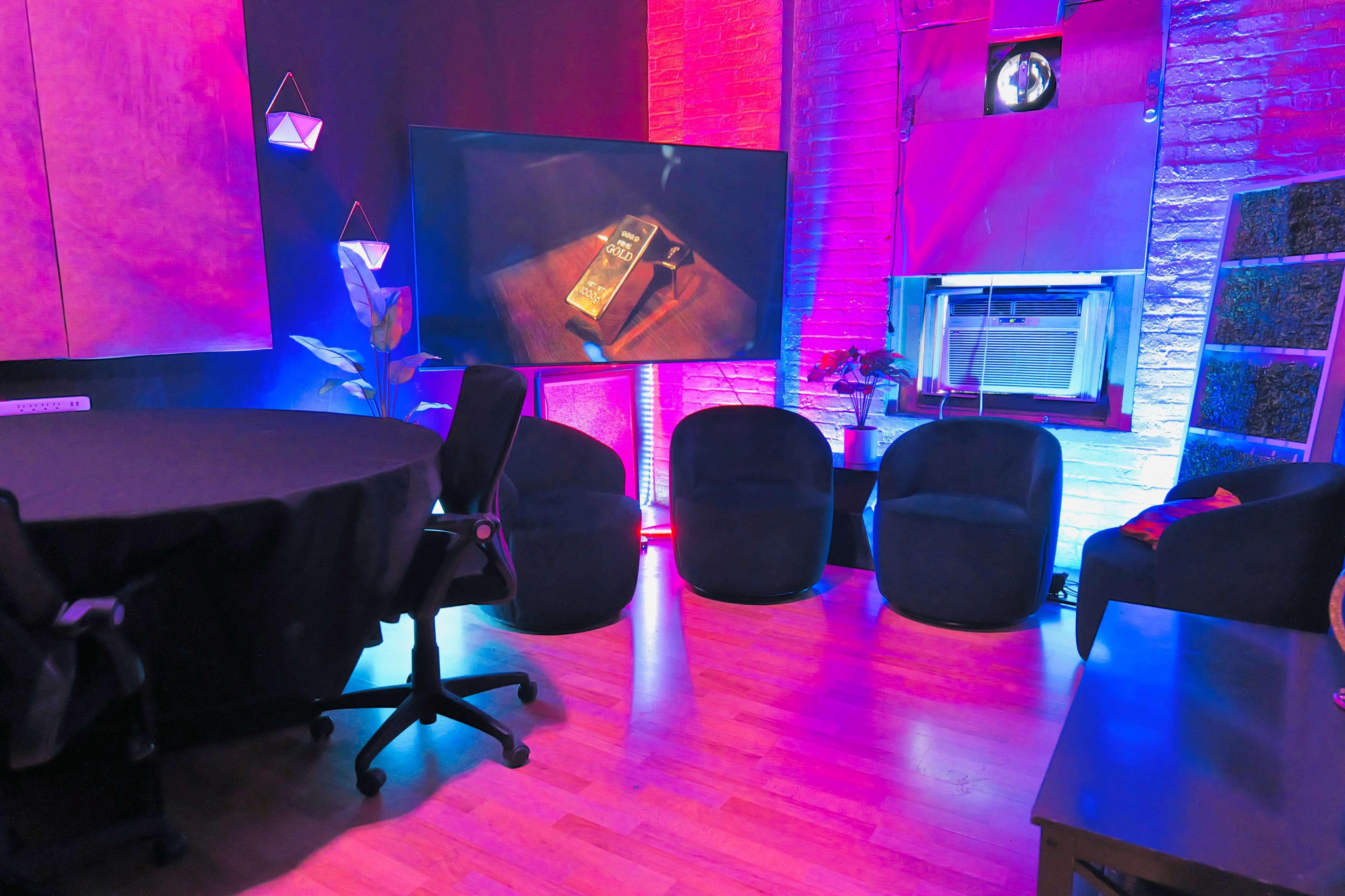 A modern indoor space featuring a round table, four black armchairs, and a large screen displaying a book, with colorful lighting and decorative elements on the walls.