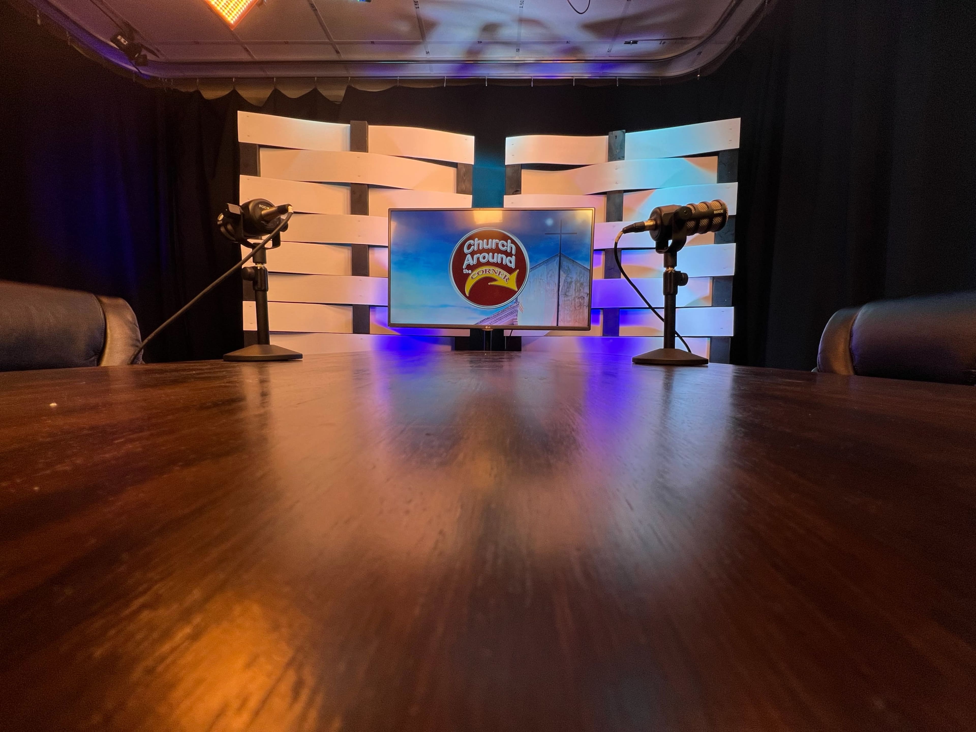 A modern studio setup with a round wooden table, two microphones, and a TV screen displaying the logo for "Church Around" against a backdrop of white panels and black curtains.