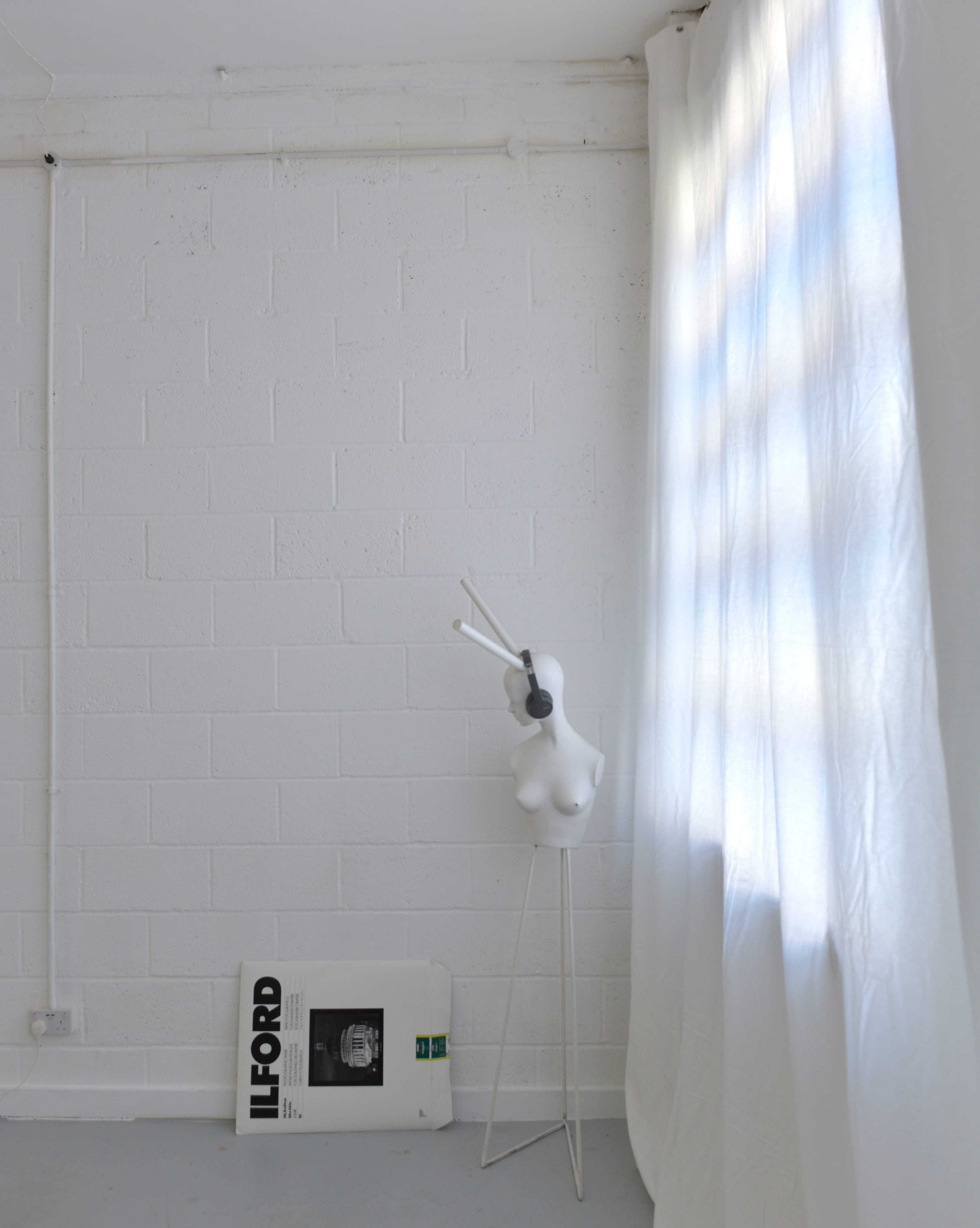 A white mannequin stands beside a folded white curtain and an Ilford poster on the floor against a textured white wall.