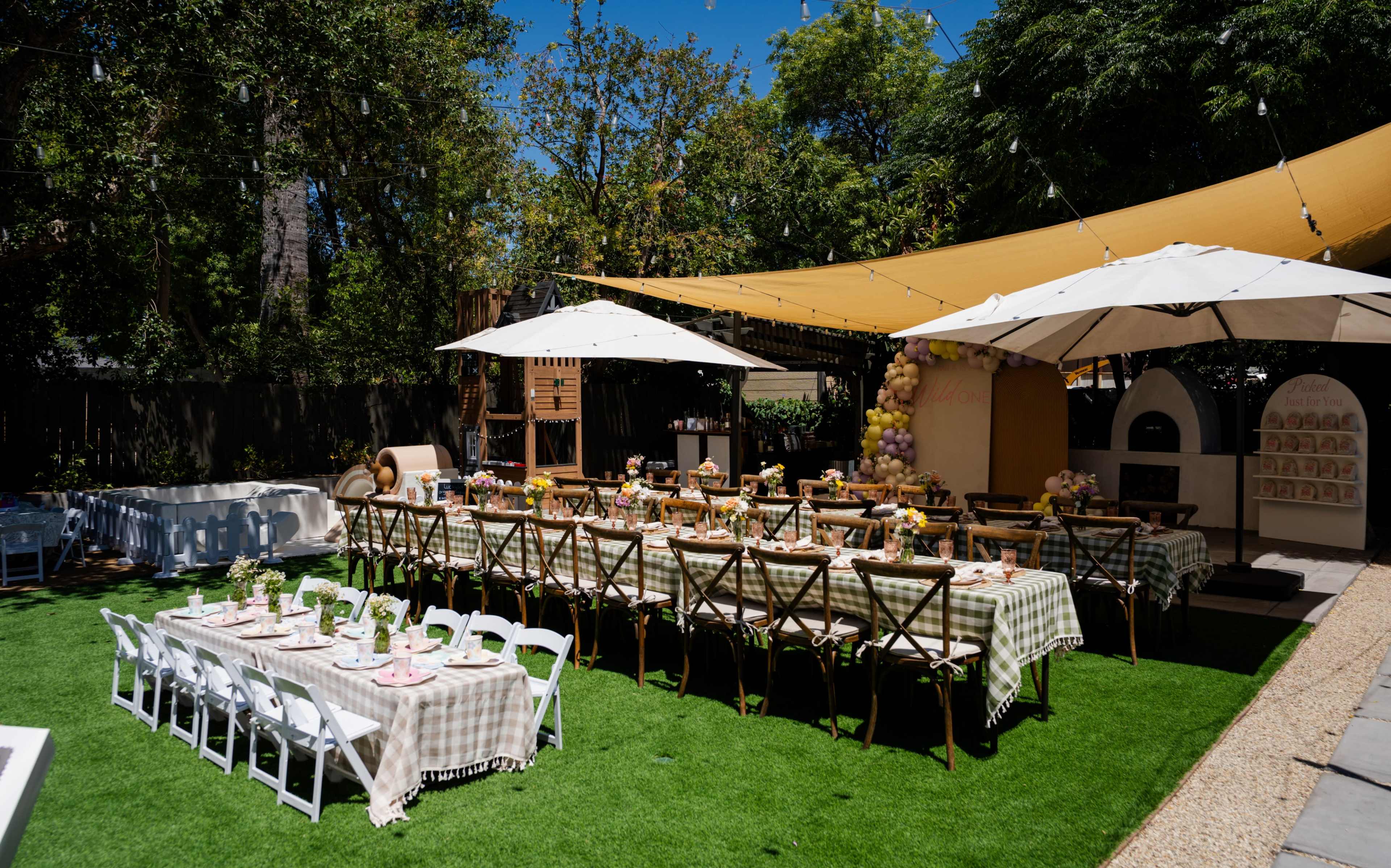 A backyard party setup features long dining tables covered with checkered tablecloths, surrounded by chairs, and shaded by large umbrellas under a sunny sky.