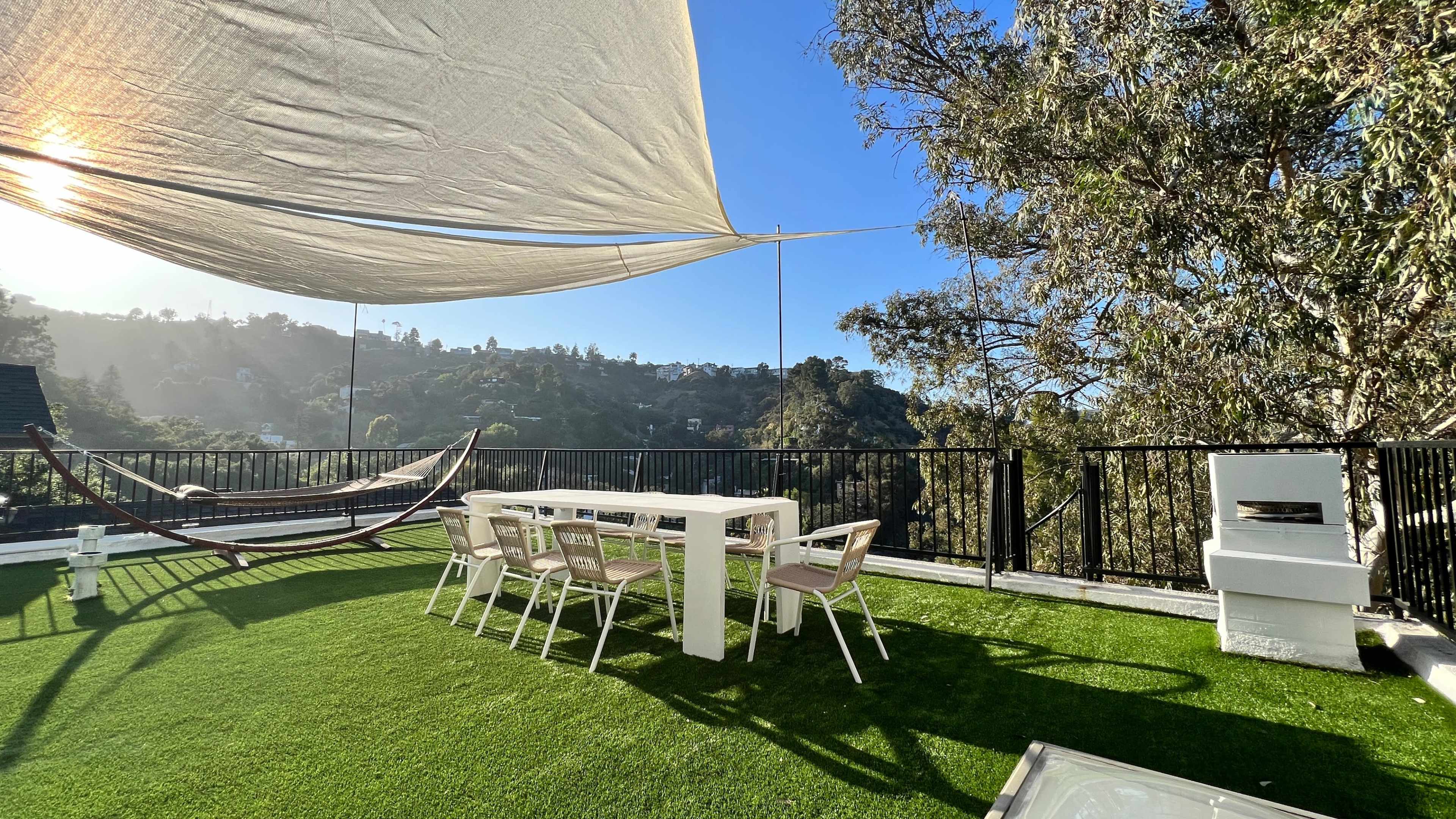 The image shows a rooftop patio with a white dining table and chairs, a hammock, and a grill, all set against a backdrop of trees and hills under a shade sail.