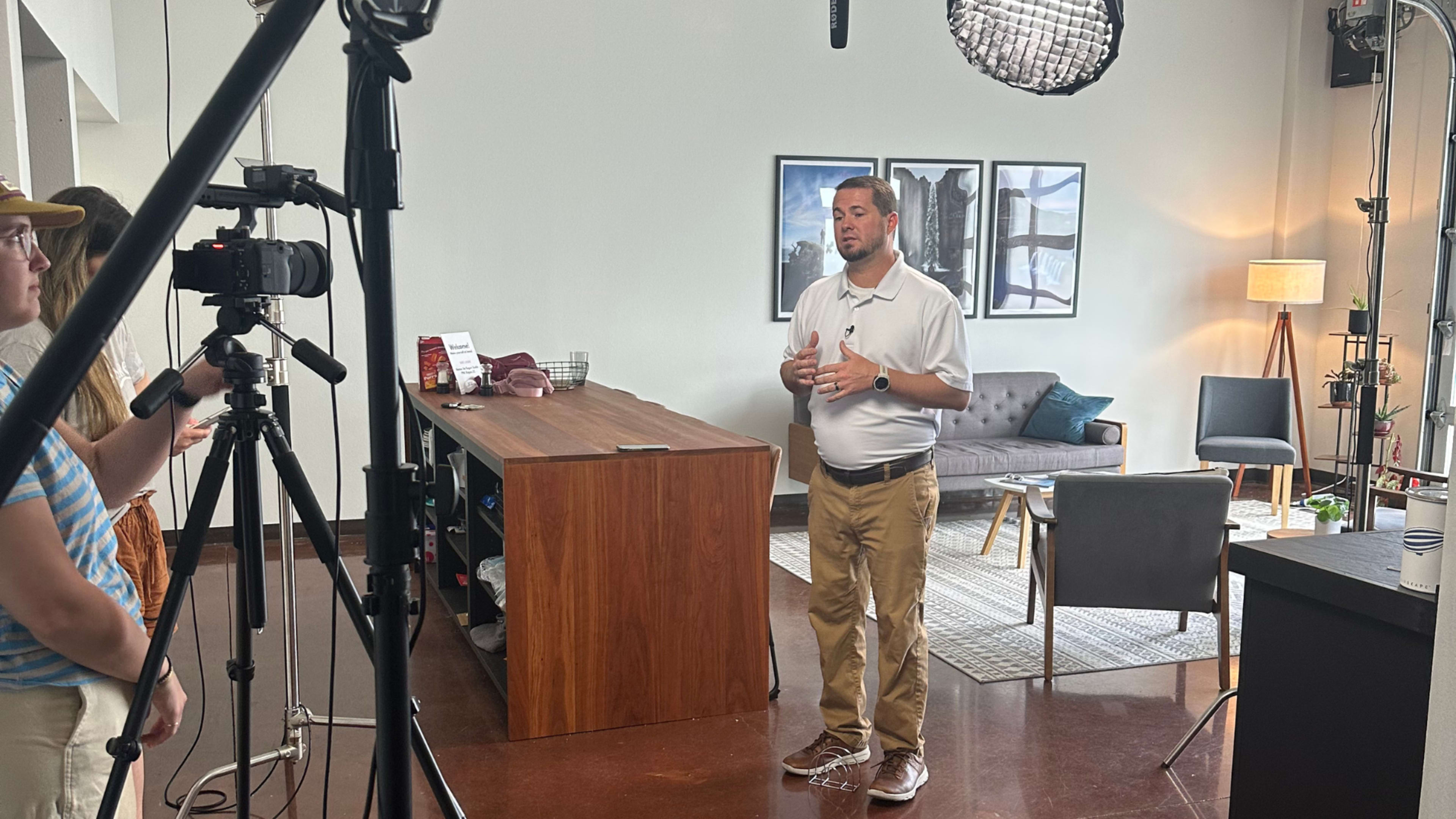 A man stands in a well-lit room, speaking into a camera set on a tripod while a second person operates the camera and equipment nearby.