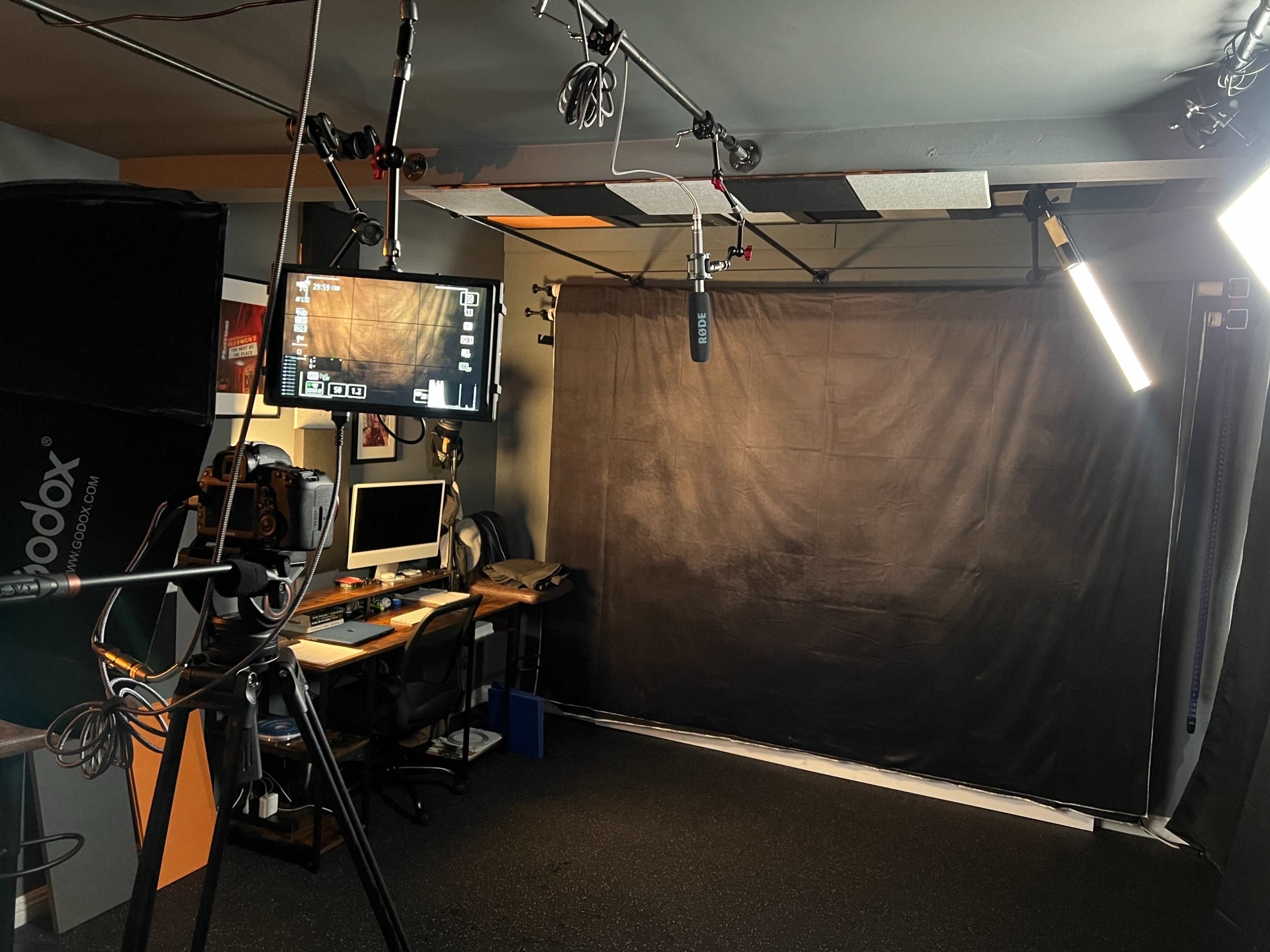 A recording studio setup featuring a black backdrop, camera equipment, microphones, a computer station, and multiple light sources.