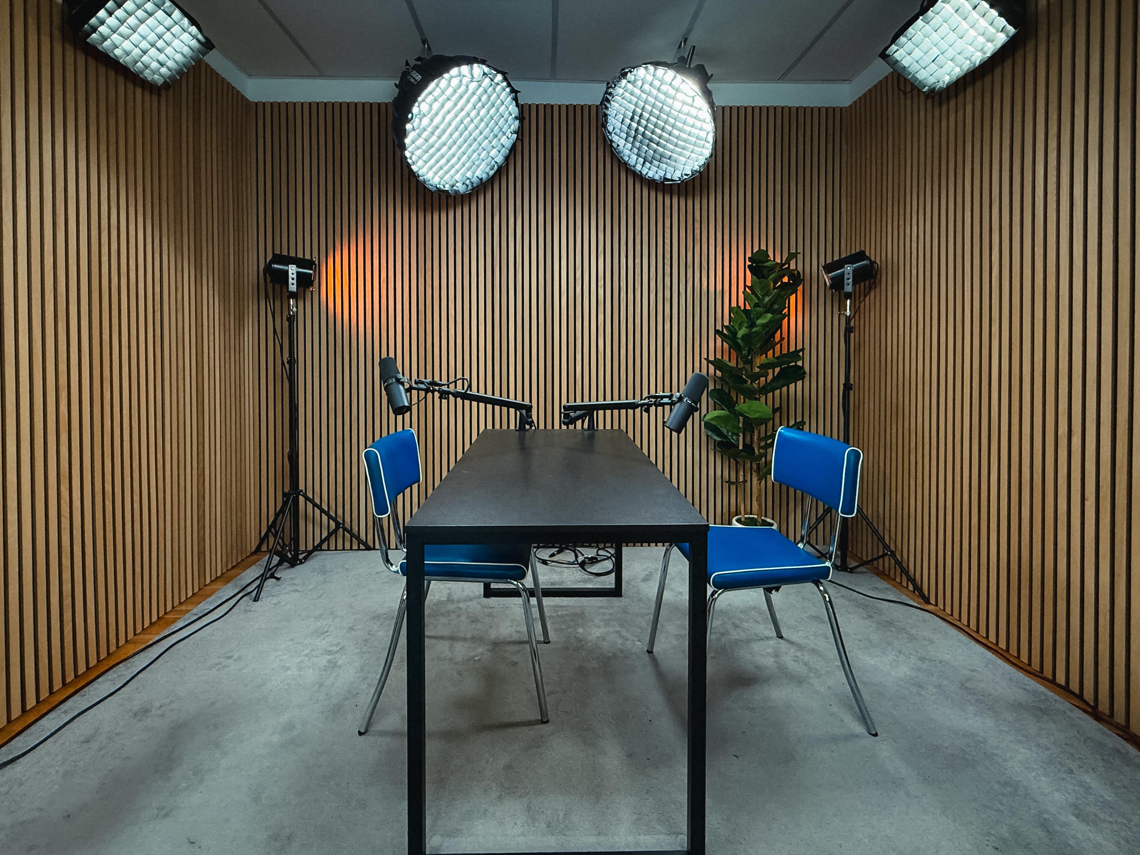 The image shows a modern recording studio with a table, two blue chairs, and several overhead lights, all set against a backdrop of wooden paneling.