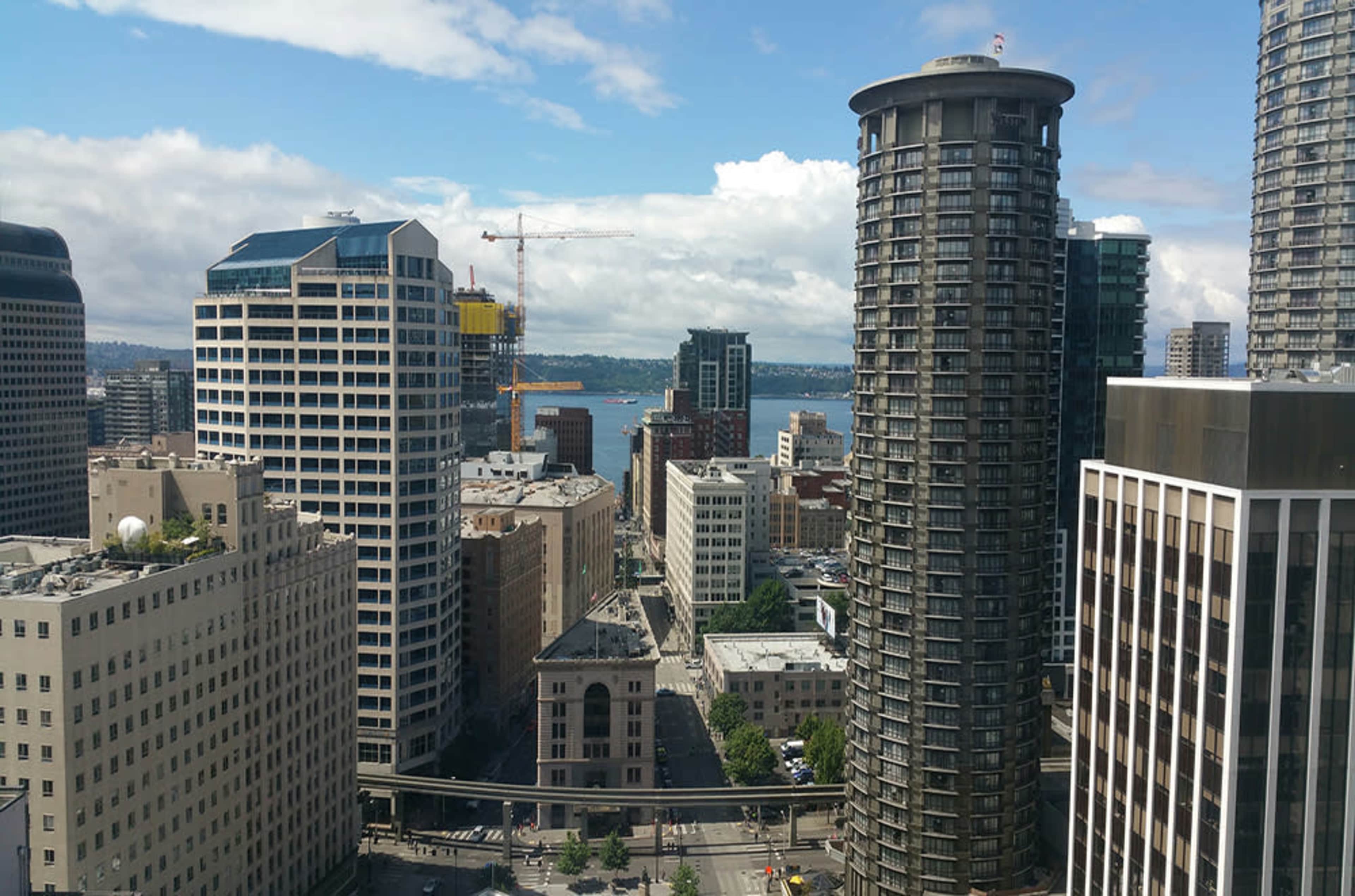 The image shows a cityscape with tall buildings, including a cylindrical skyscraper, and a view of a body of water in the background under a partly cloudy sky.