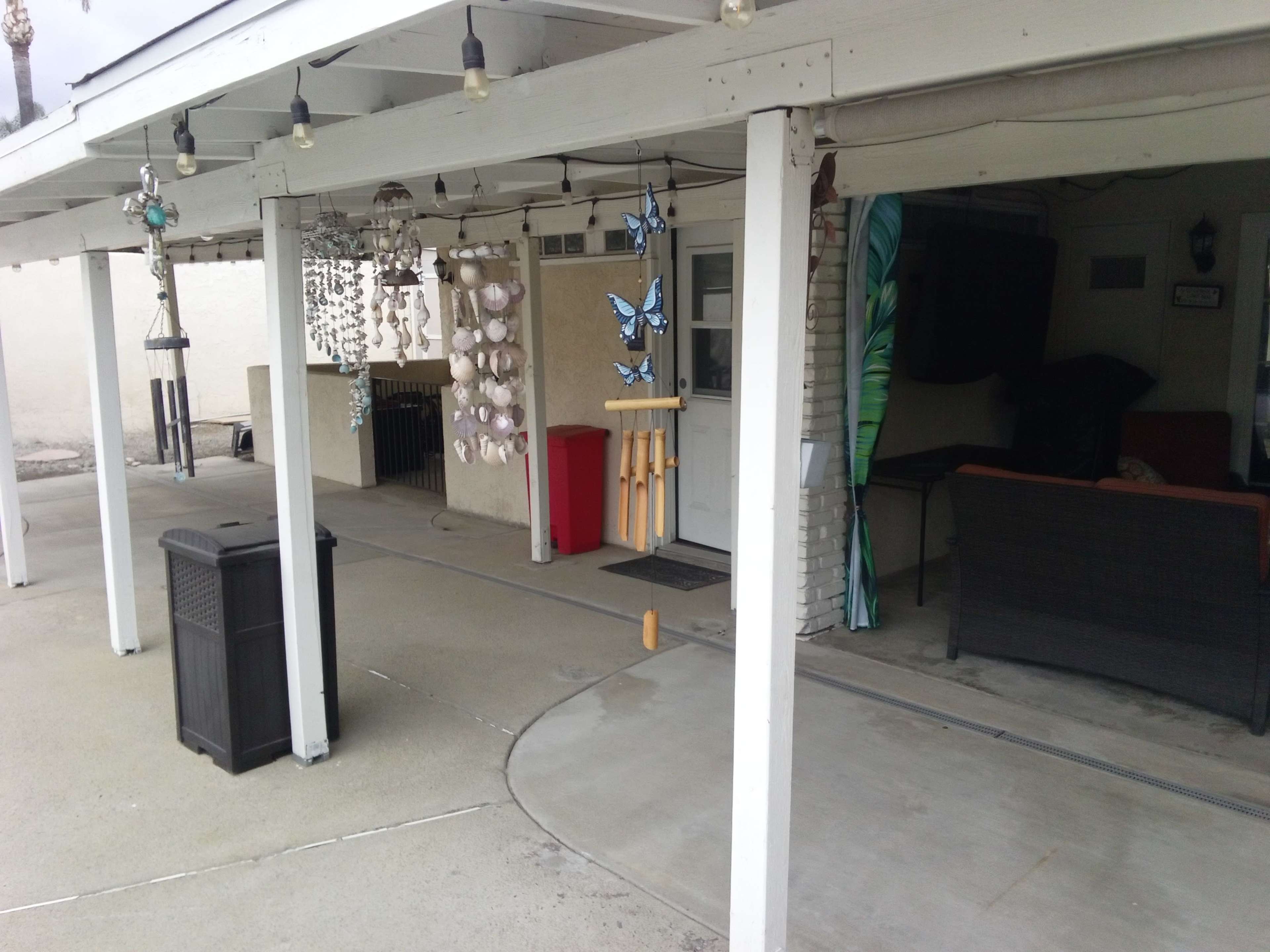 The image shows a covered patio area with hanging wind chimes, a red storage bin, and a seating arrangement against the wall.