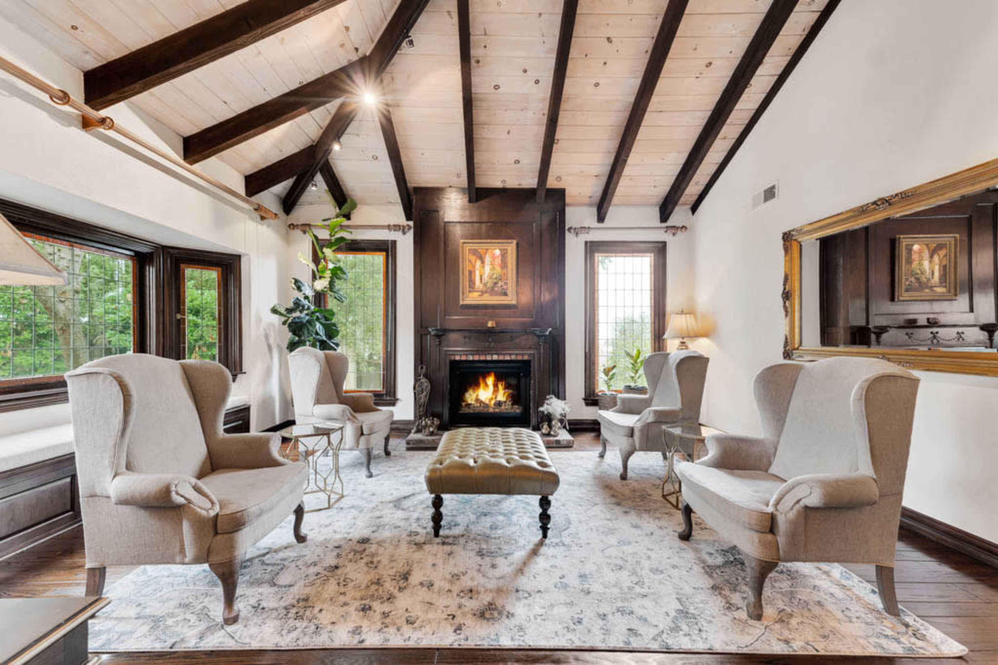 The living room features a central fireplace, surrounded by four upholstered chairs and a tufted ottoman, with wooden beams accentuating the ceiling.