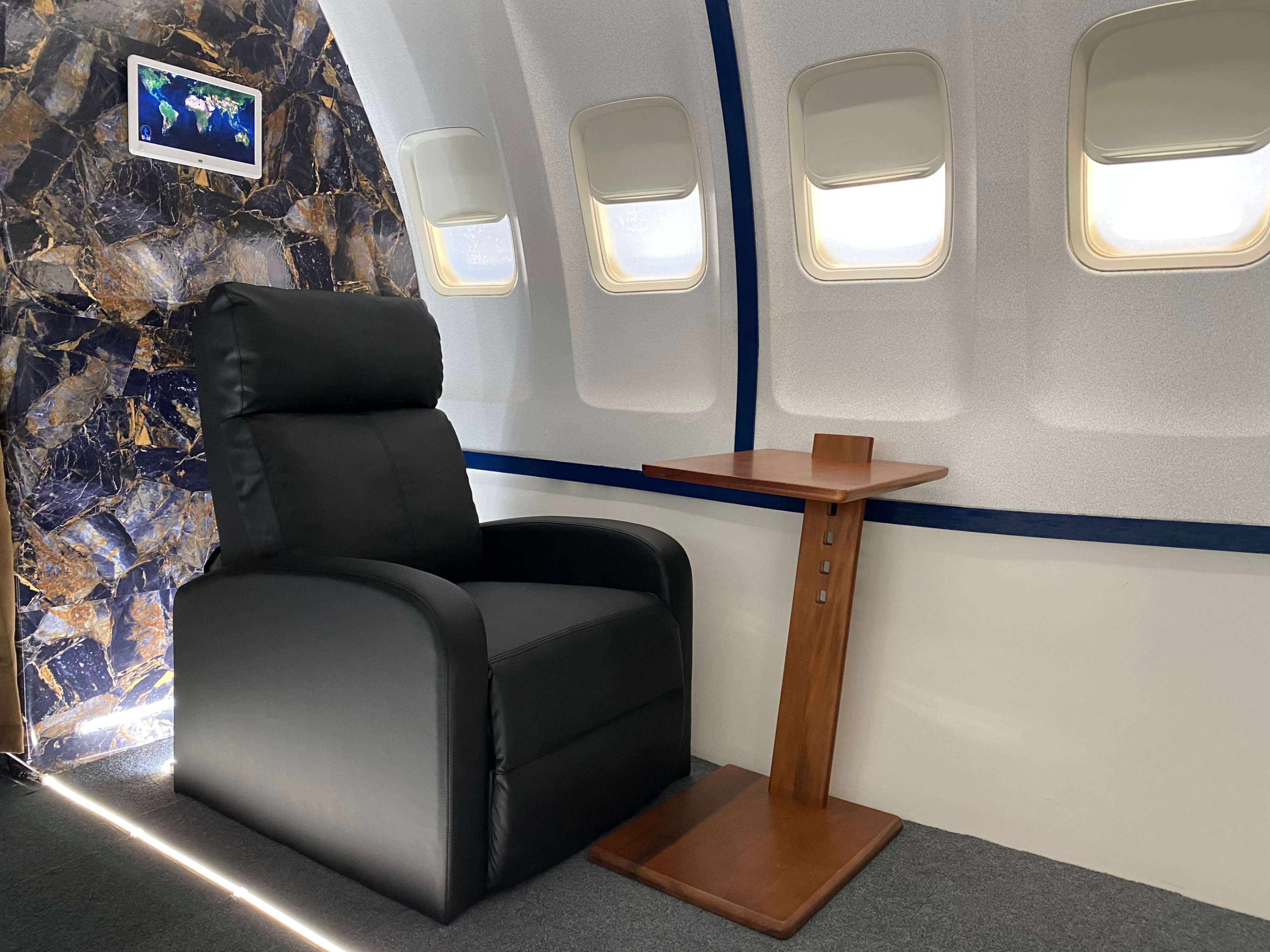 A black recliner chair next to a wooden side table inside the cabin of an aircraft, with a framed map hanging on the wall.