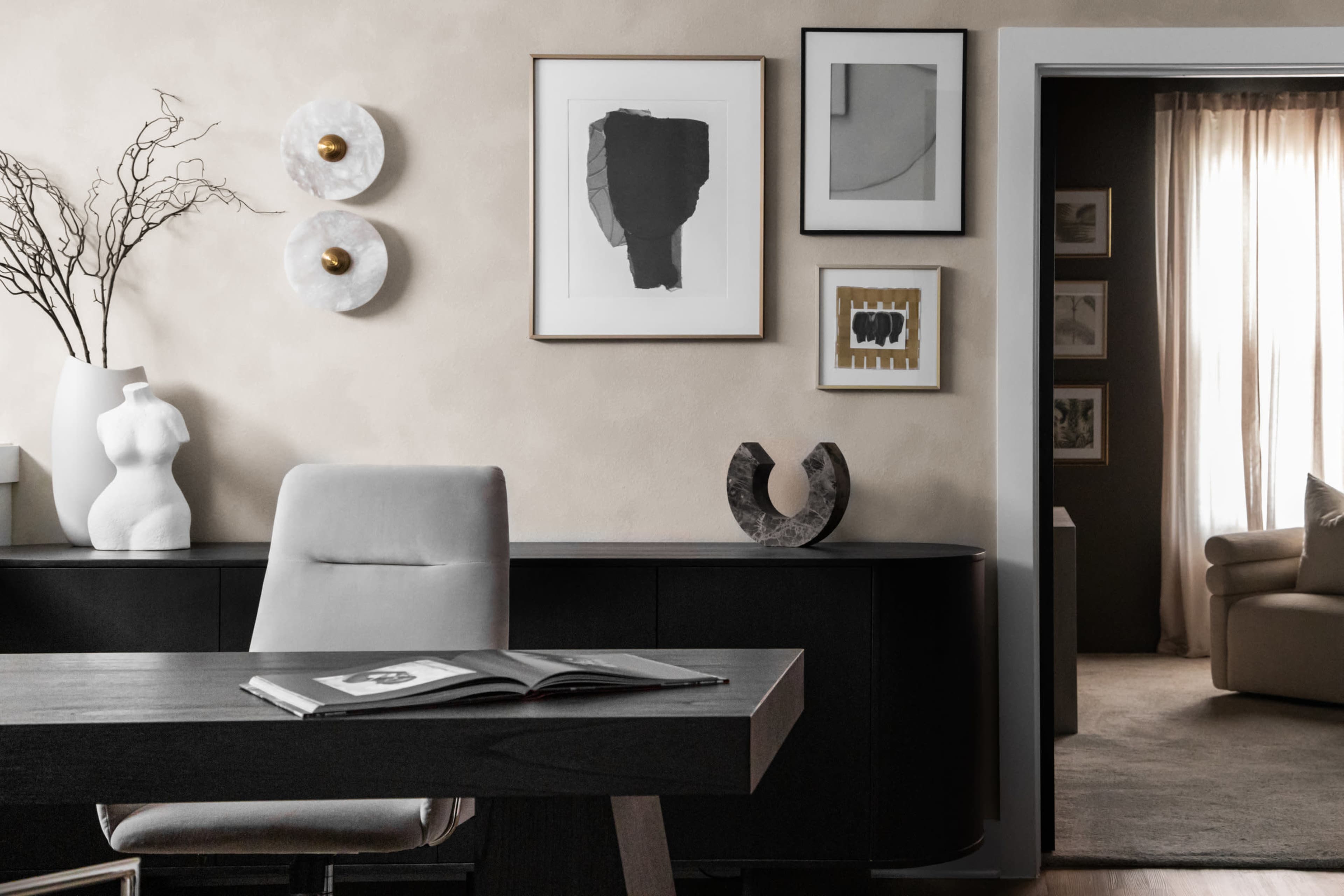 The image shows a modern office space featuring a sleek black desk, decorative wall art, a sculptural white figure, and a softly lit doorway leading to a living area.