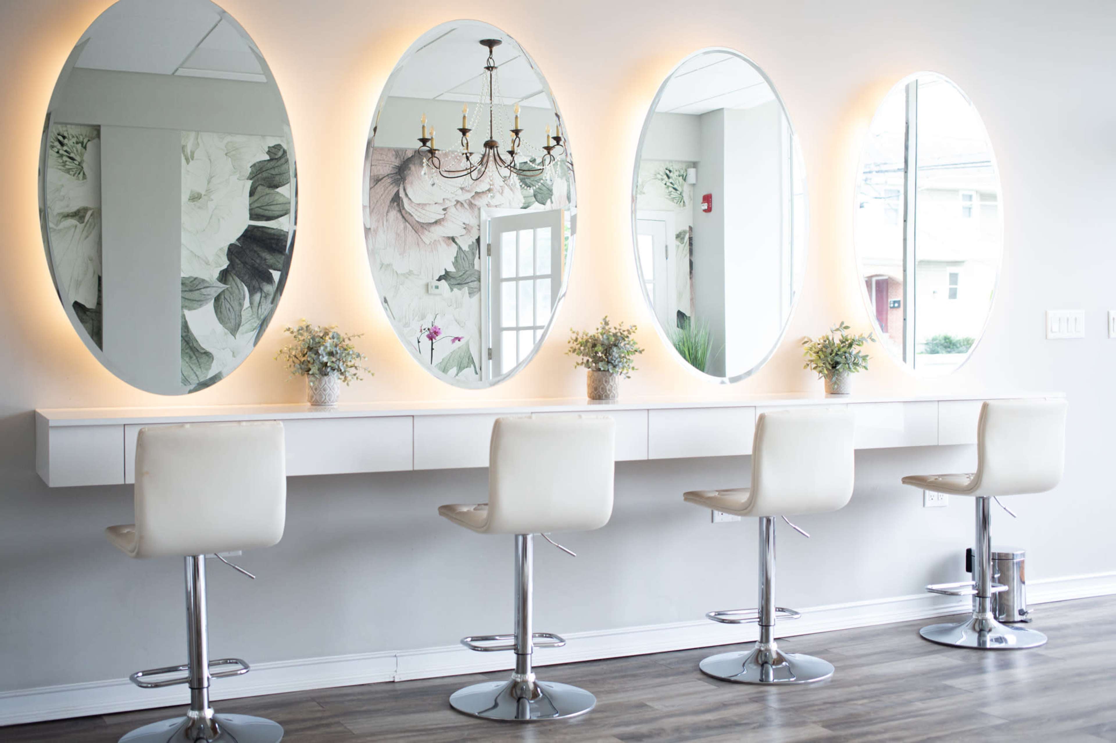 A modern vanity area with four white bar stools positioned in front of large oval mirrors illuminated by soft lights.
