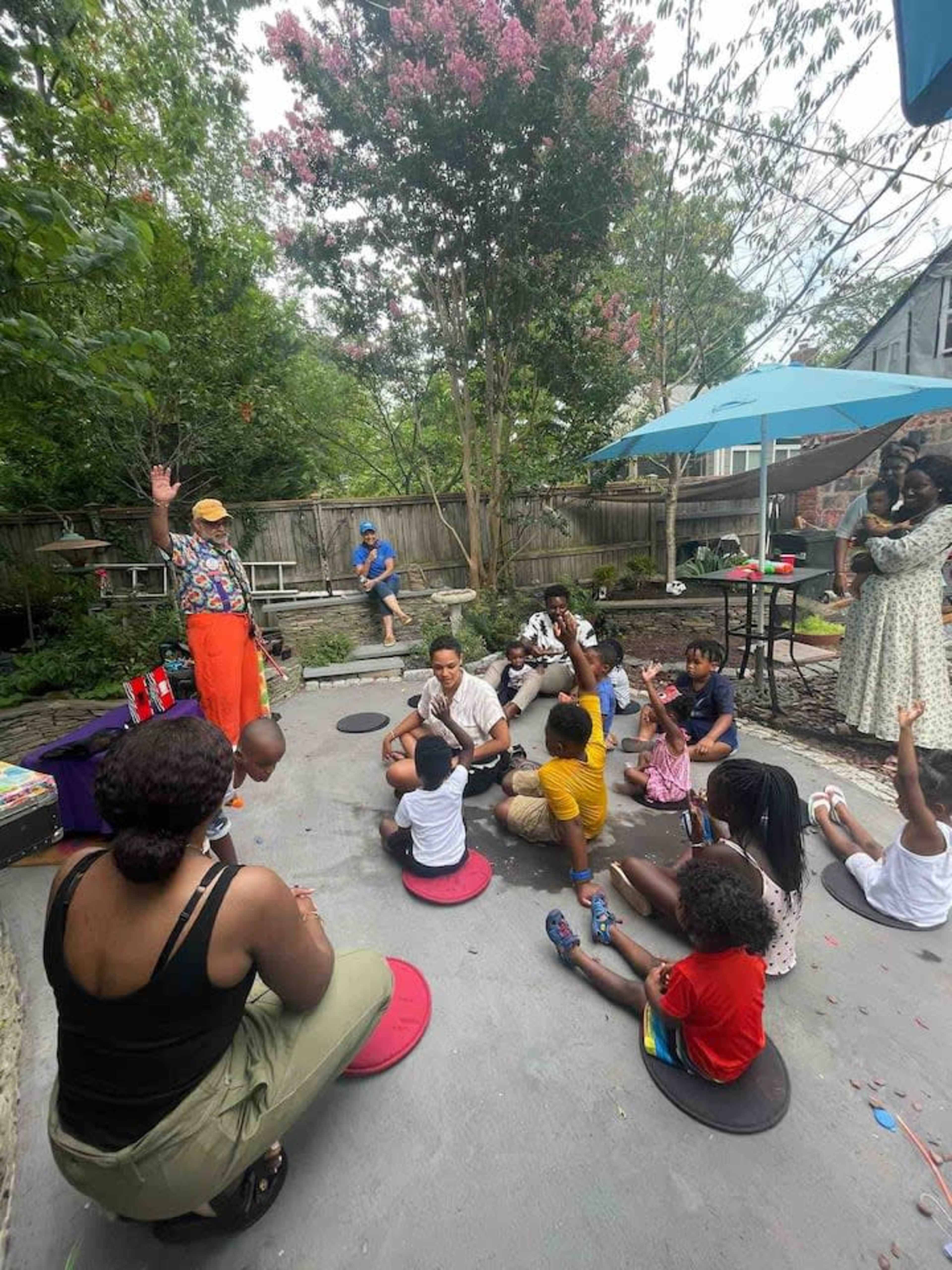 A group of children is sitting on circular mats in a backyard while an entertainer engages them, with adults observing nearby under an umbrella.