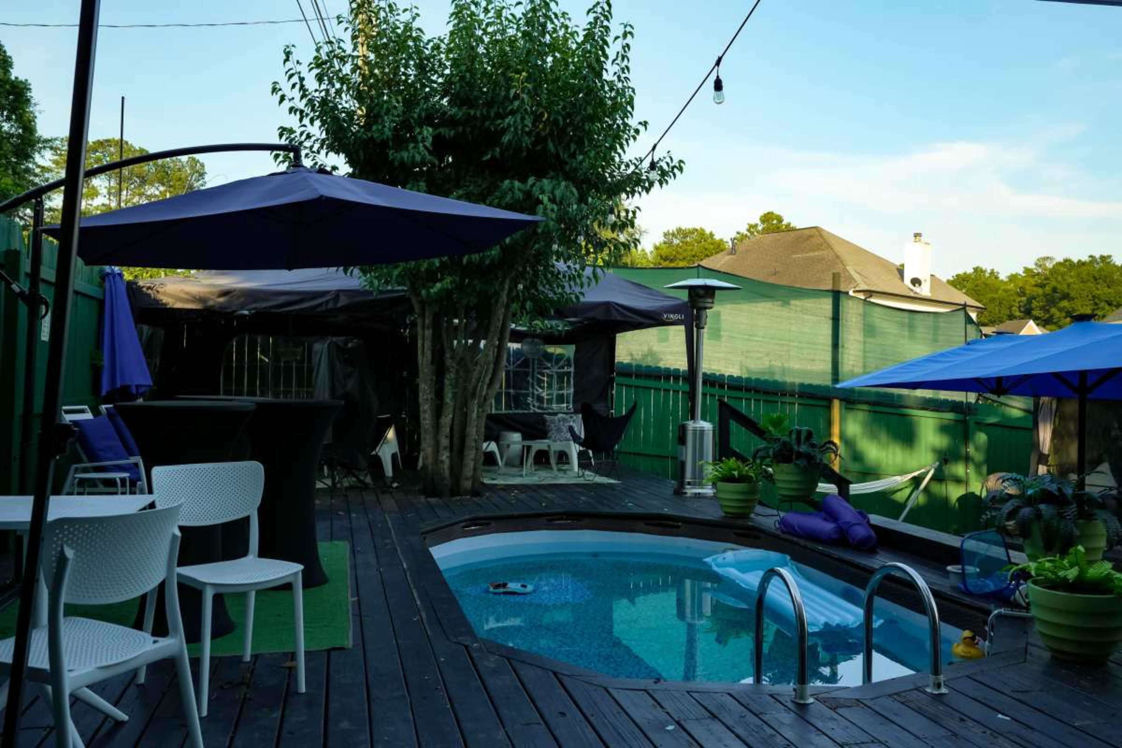 The image features a backyard space with a small swimming pool surrounded by wooden decking, patio furniture, and various plants under clear skies.