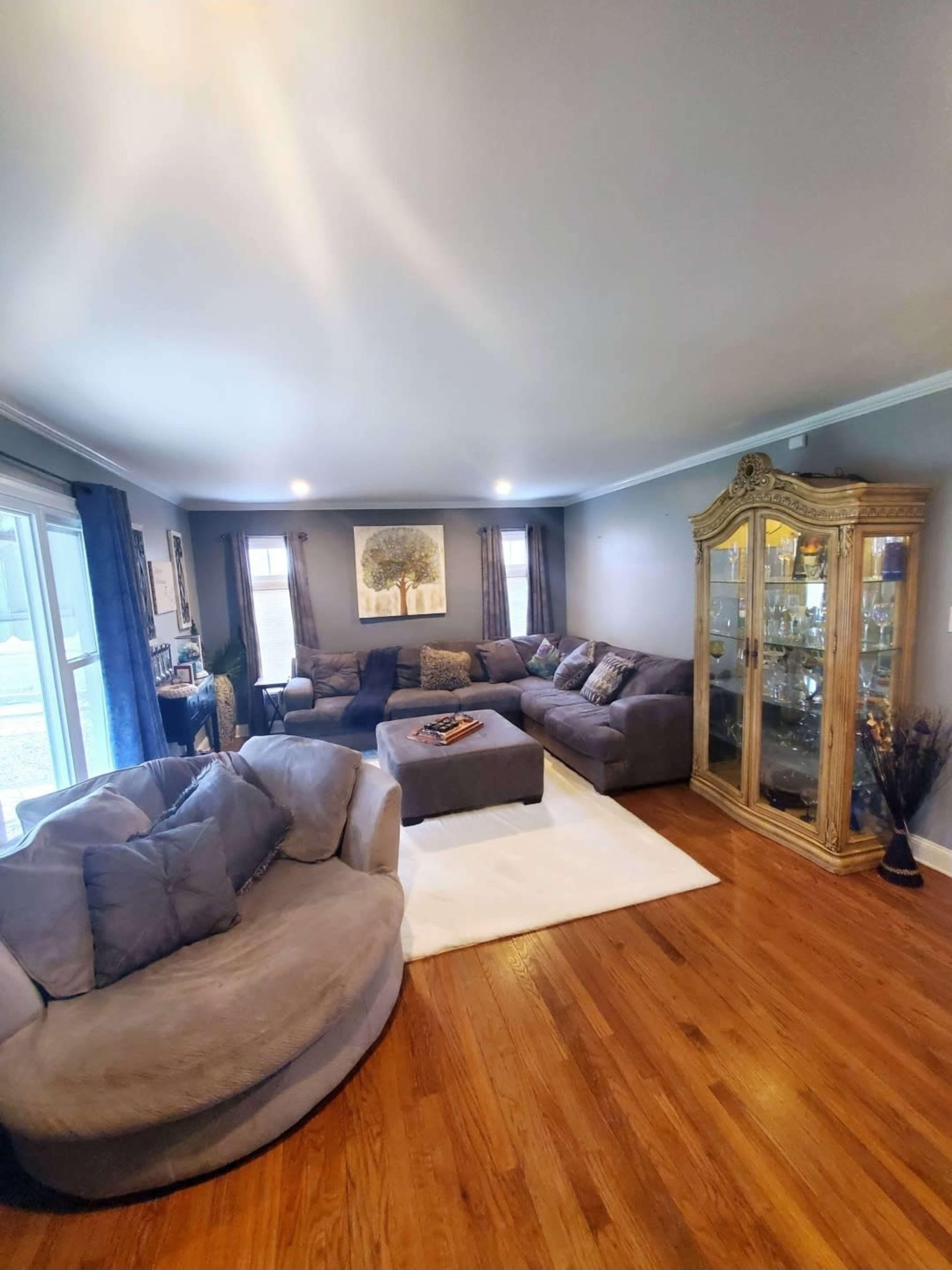 The image shows a living room with a large sectional sofa and a glass-fronted cabinet, featuring hardwood flooring and natural light coming through the windows.