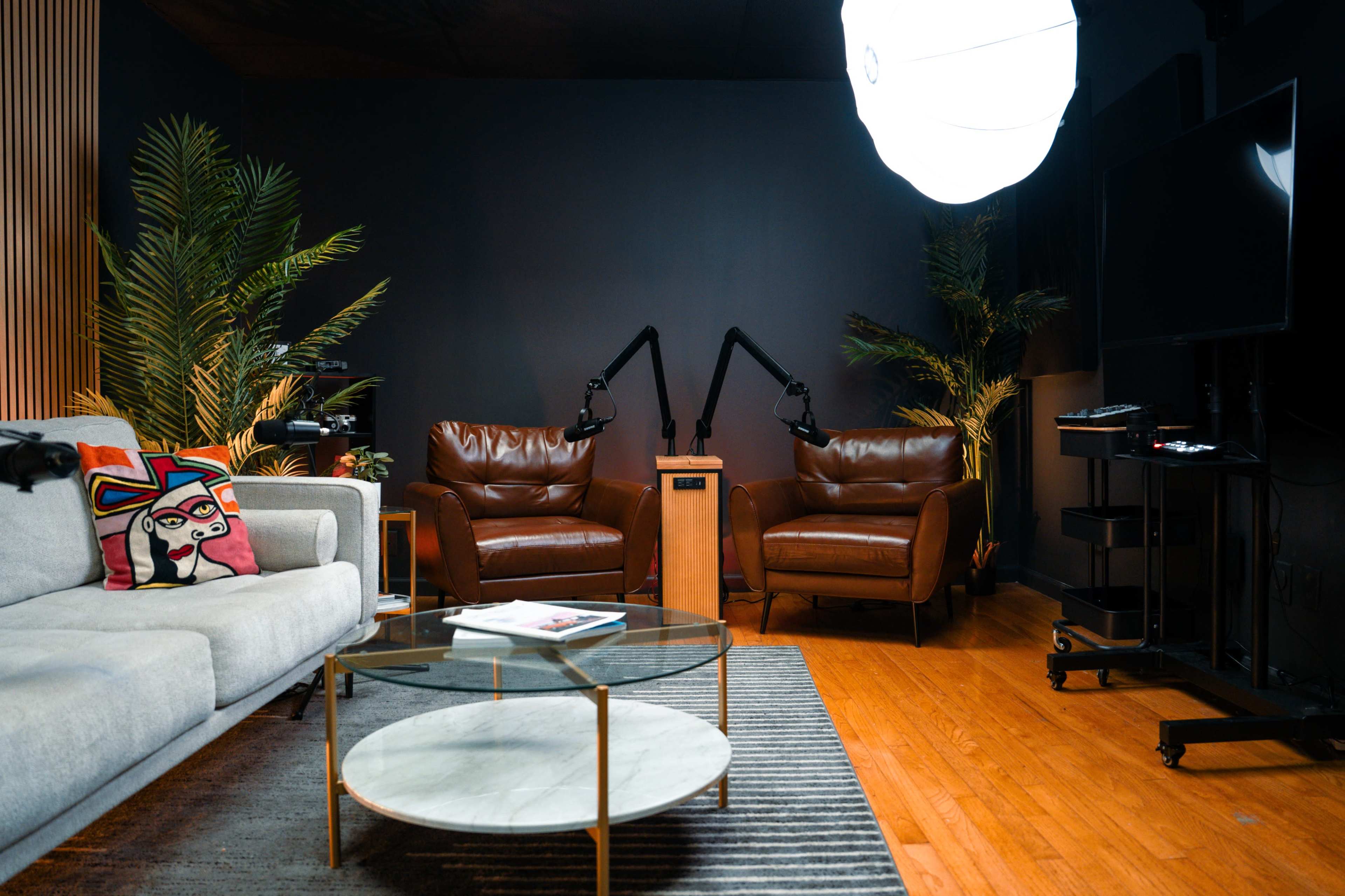 The image shows a modern, dimly lit living space with a gray couch, two brown leather chairs, a round glass coffee table, and a large light source overhead.