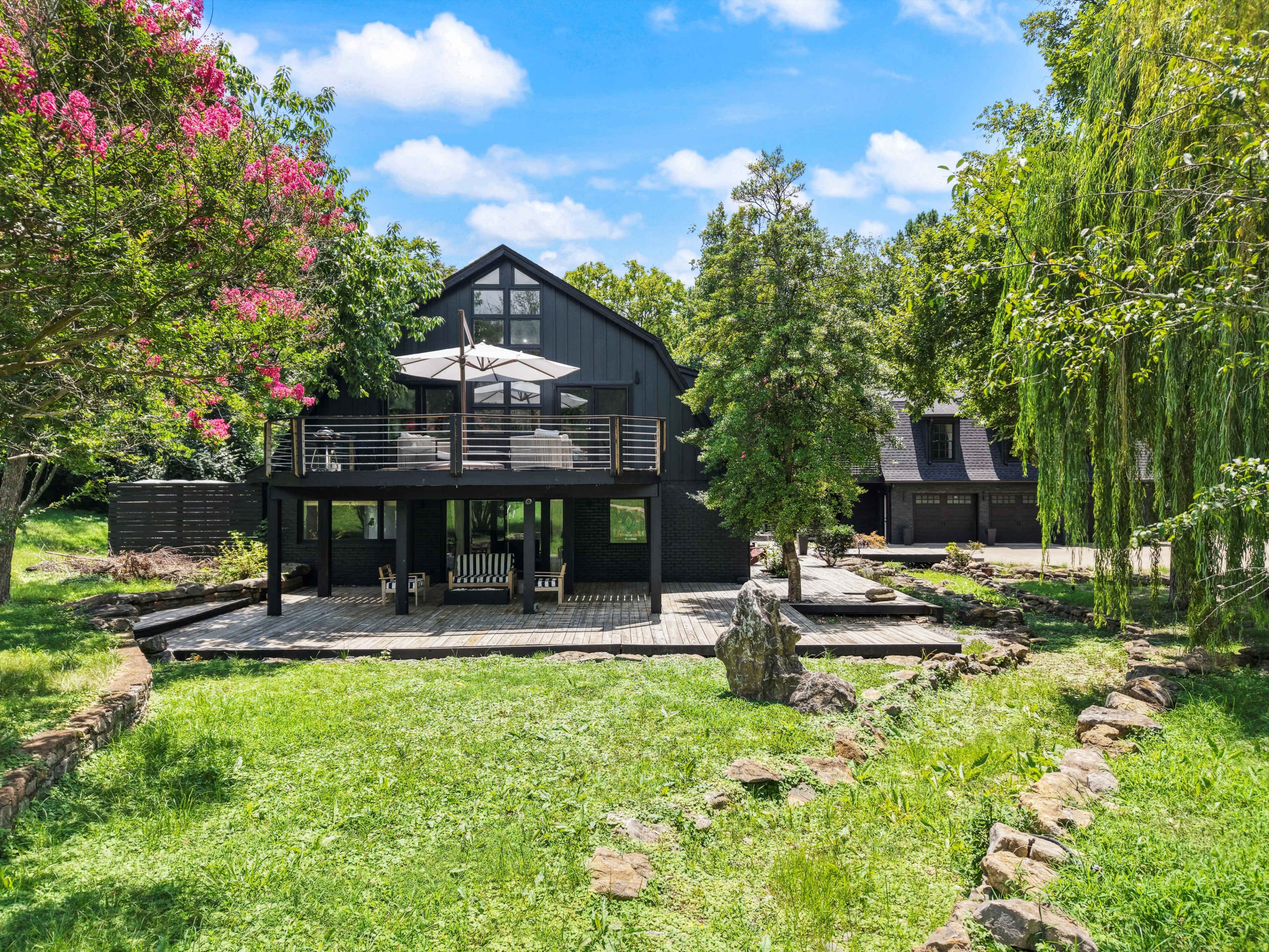 The image shows a modern black house surrounded by greenery, featuring a deck with outdoor seating and a nearby garage.