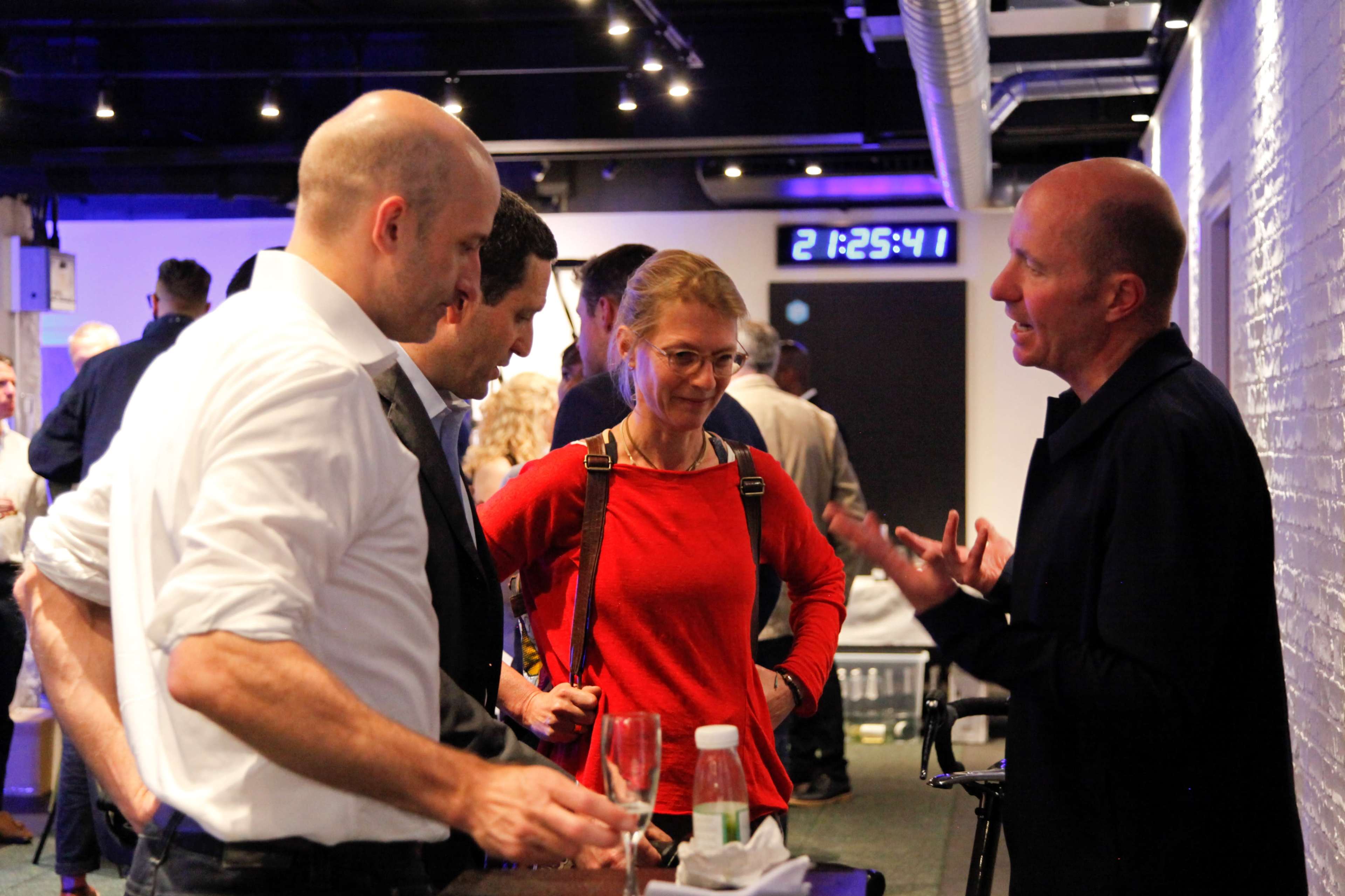 Four people engage in conversation at a darkly lit event, with one person speaking animatedly while others listen attentively.