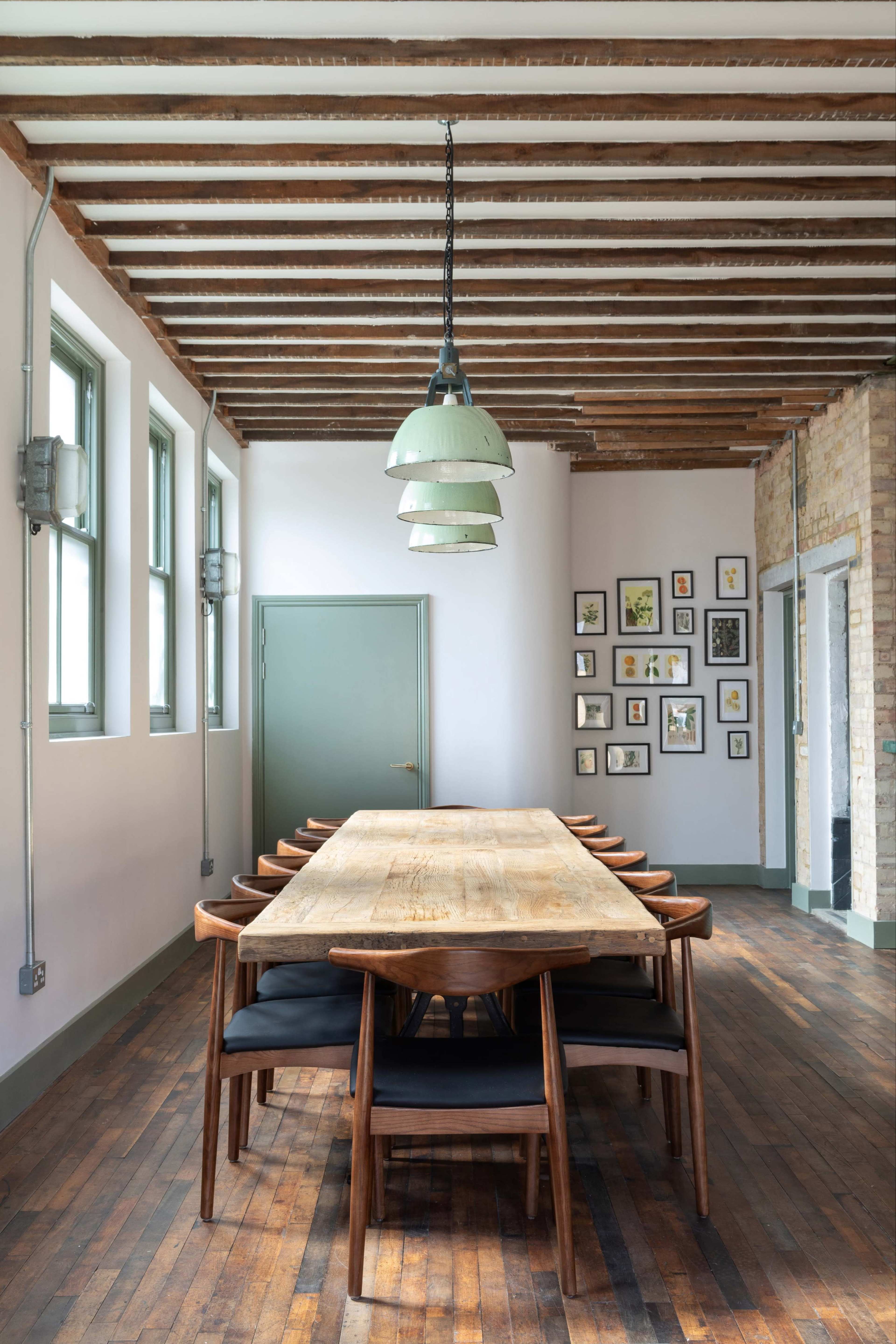 A long wooden dining table with seated chairs occupies the center of a room featuring exposed beams, a green pendant light, and wall decorations.