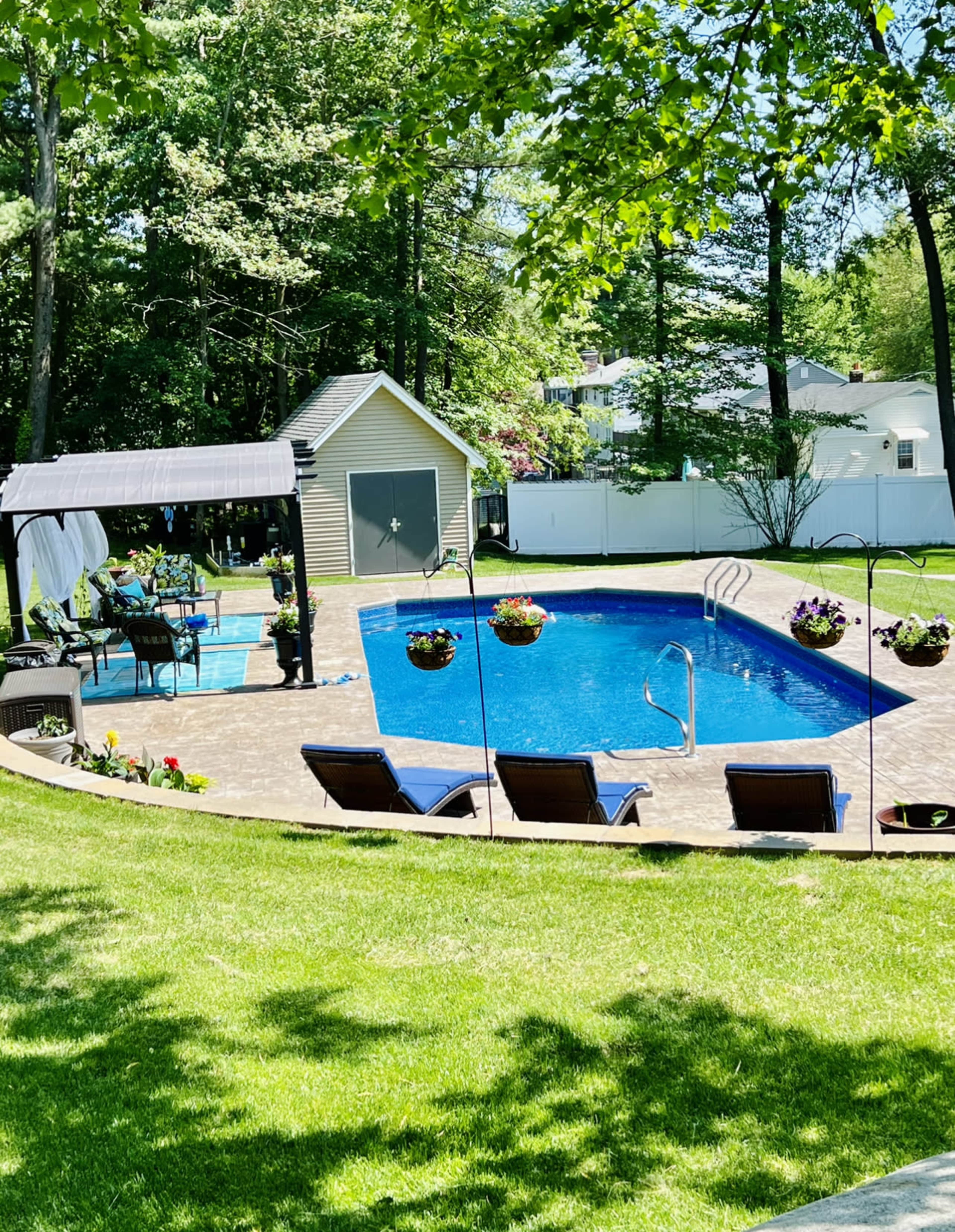 The image shows a backyard with a blue swimming pool, lounge chairs, a gazebo, and a small shed, surrounded by trees and a white fence.
