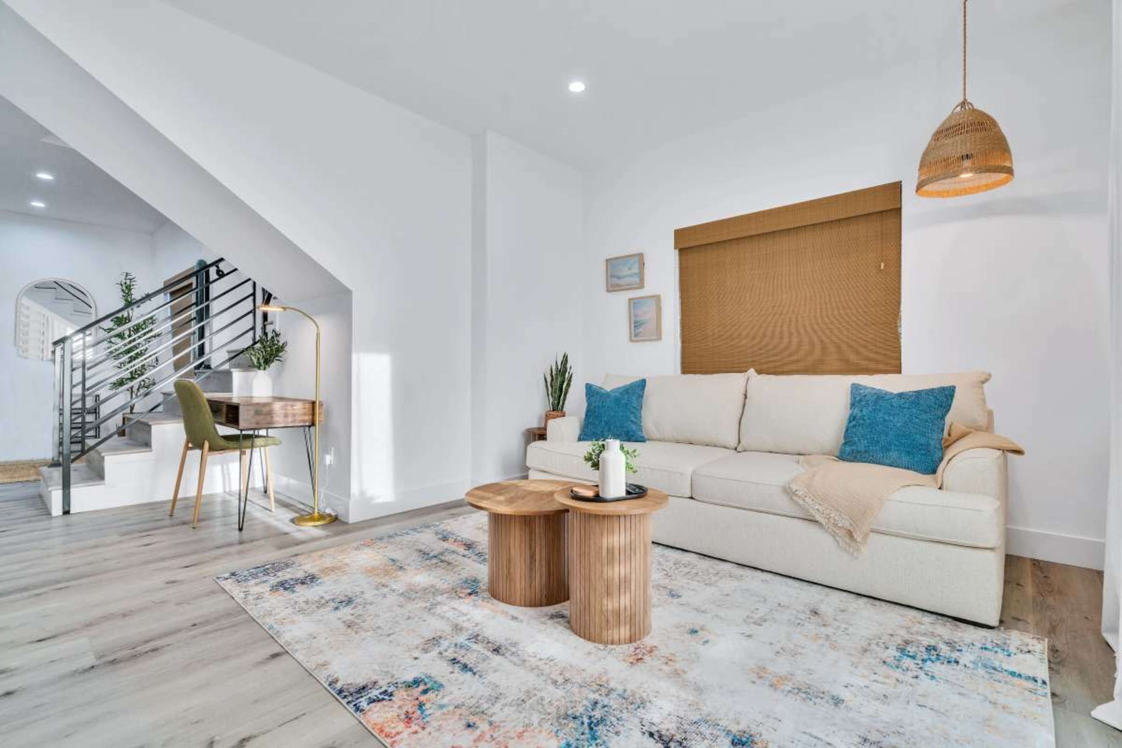 The scene features a modern living room with a cream-colored sofa, two wooden coffee tables, and a decorative rug, along with a staircase and a small workspace in the background.